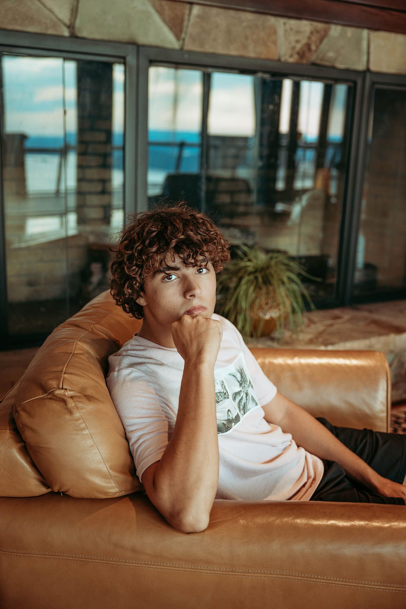 Senior in a white t-shirt reclines on a brown leather couch by large windows in low-lit interior scene.
