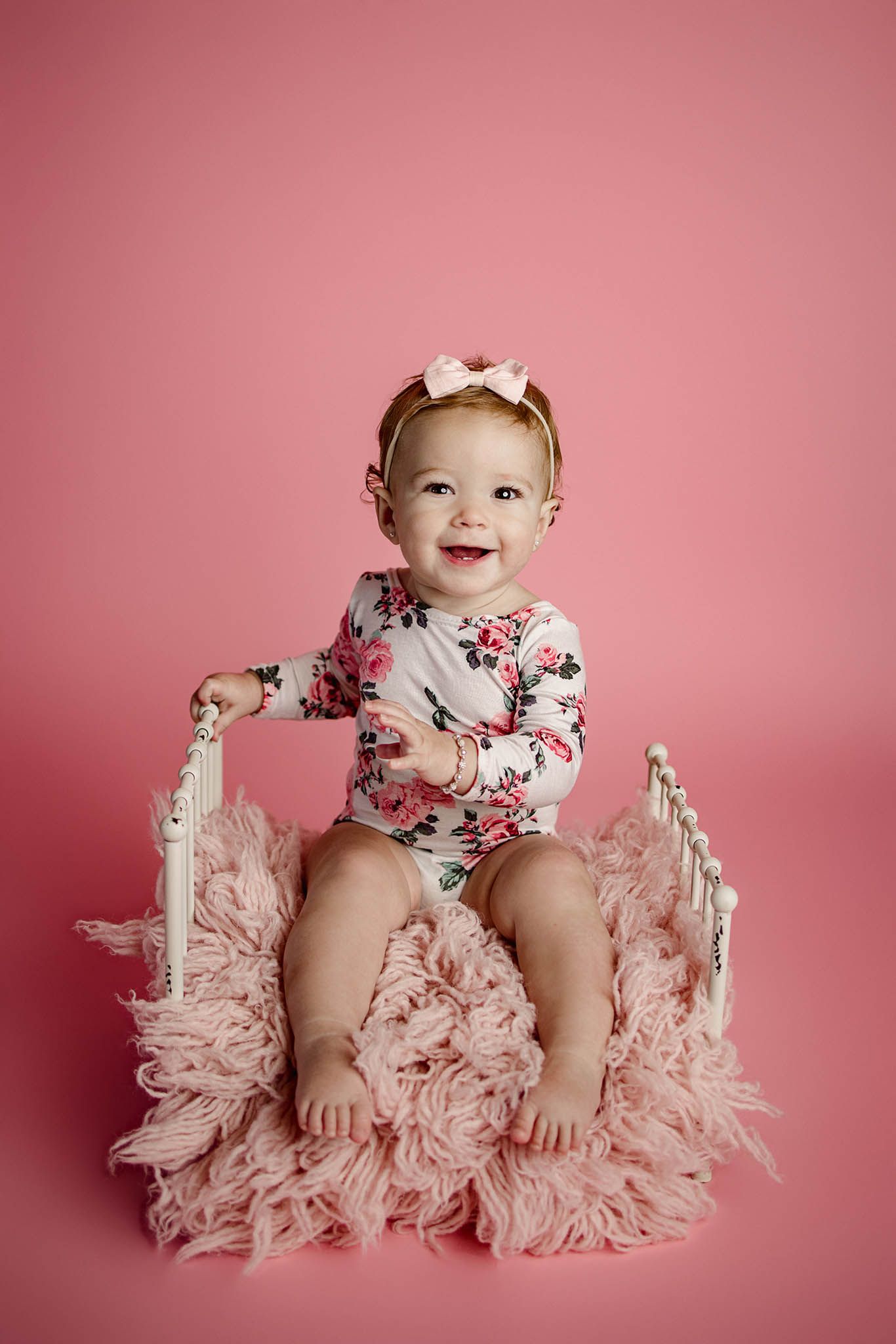 Baby in floral dress sitting on pink fluffy rug against pink backdrop.
