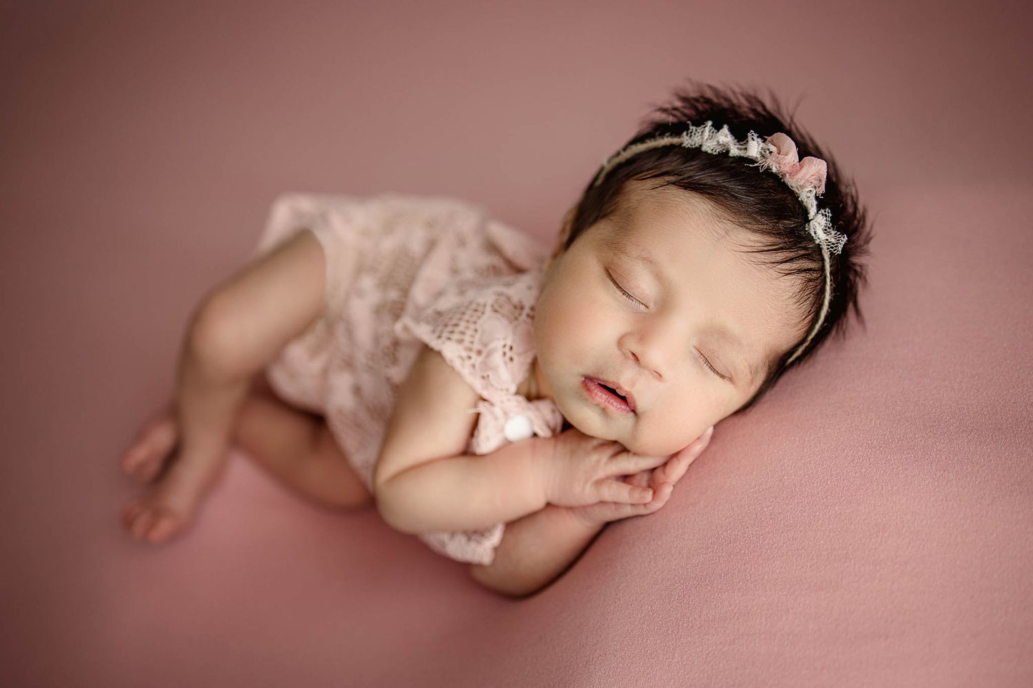 Newborn in pink lace wrap posed against rosy pink backdrop in artistic portrait.