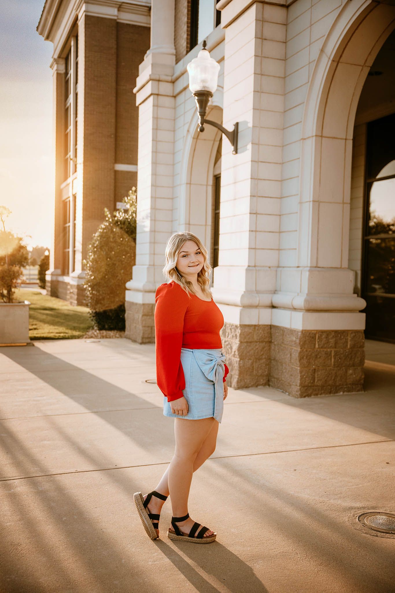 A senior wearing a bright red top and white shorts walks past white stone columns in warm sunlight.