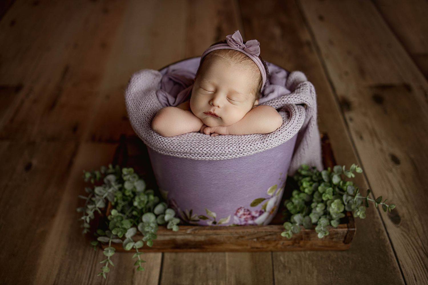 Peaceful newborn resting in purple bowl on rustic wooden floor with natural styling.