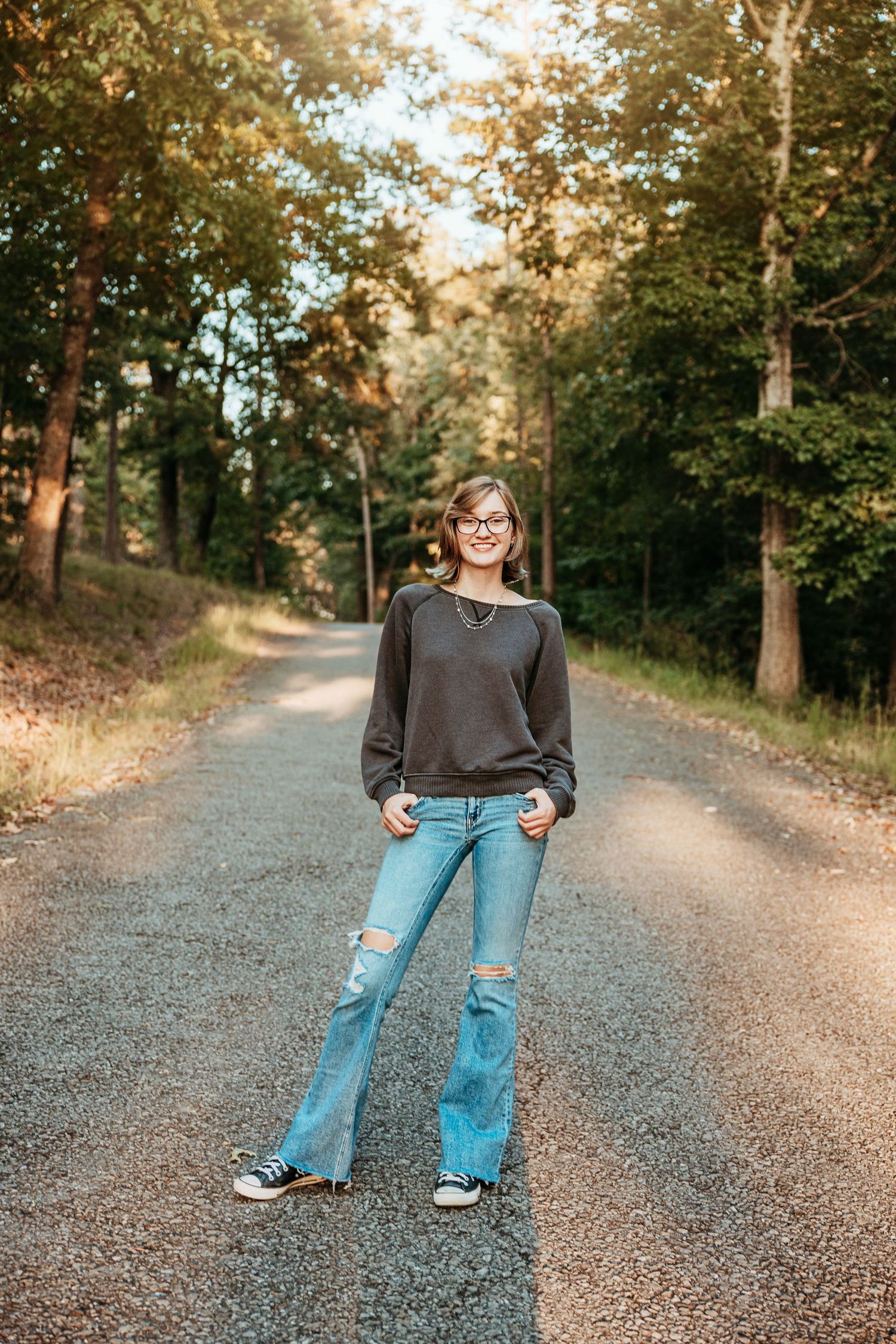 Young woman in ripped jeans and gray sweatshirt smiling on a tree-lined road during golden hour.