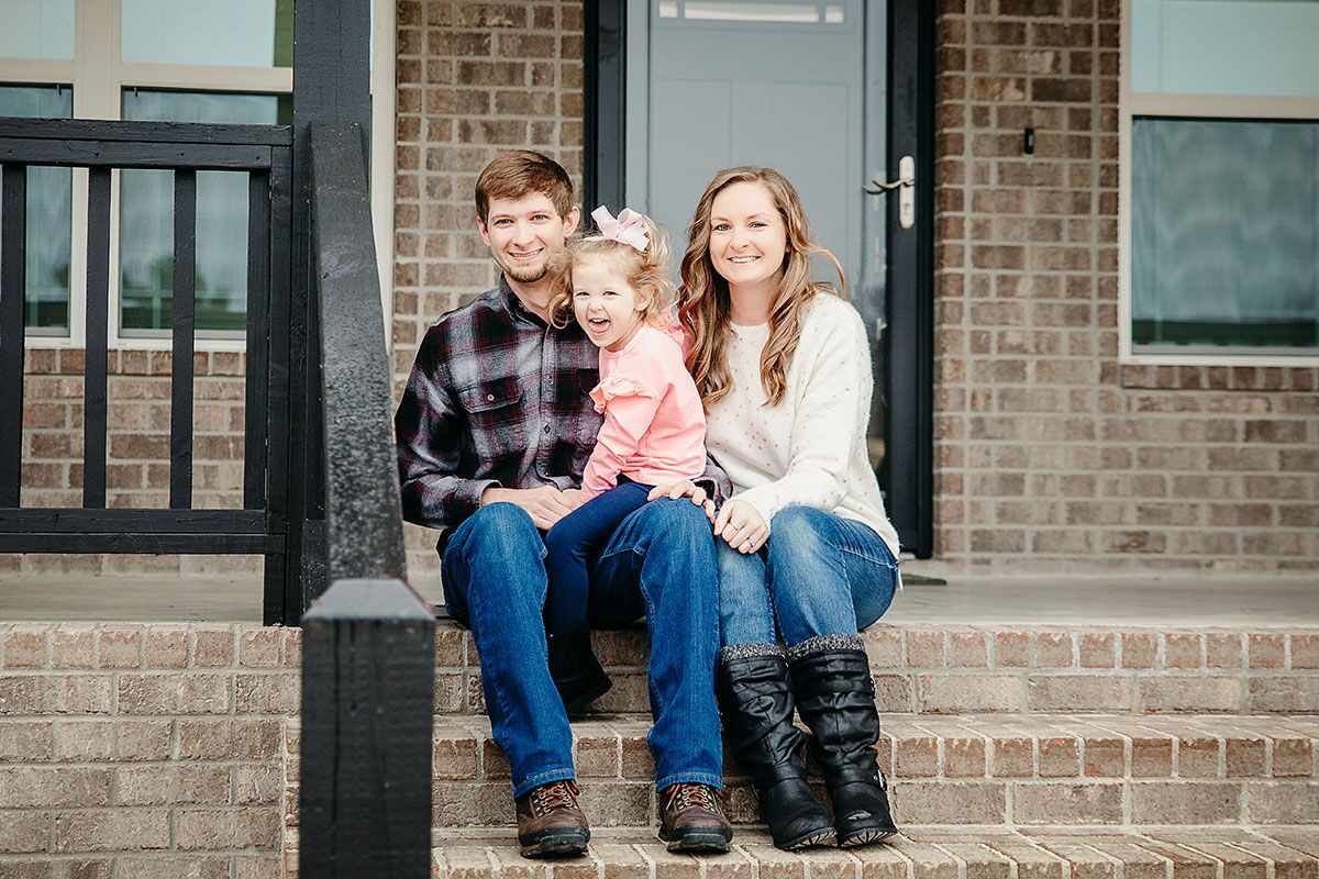 Three people sit together on brick steps outside a building entrance wearing casual clothing and smiling at the camera.