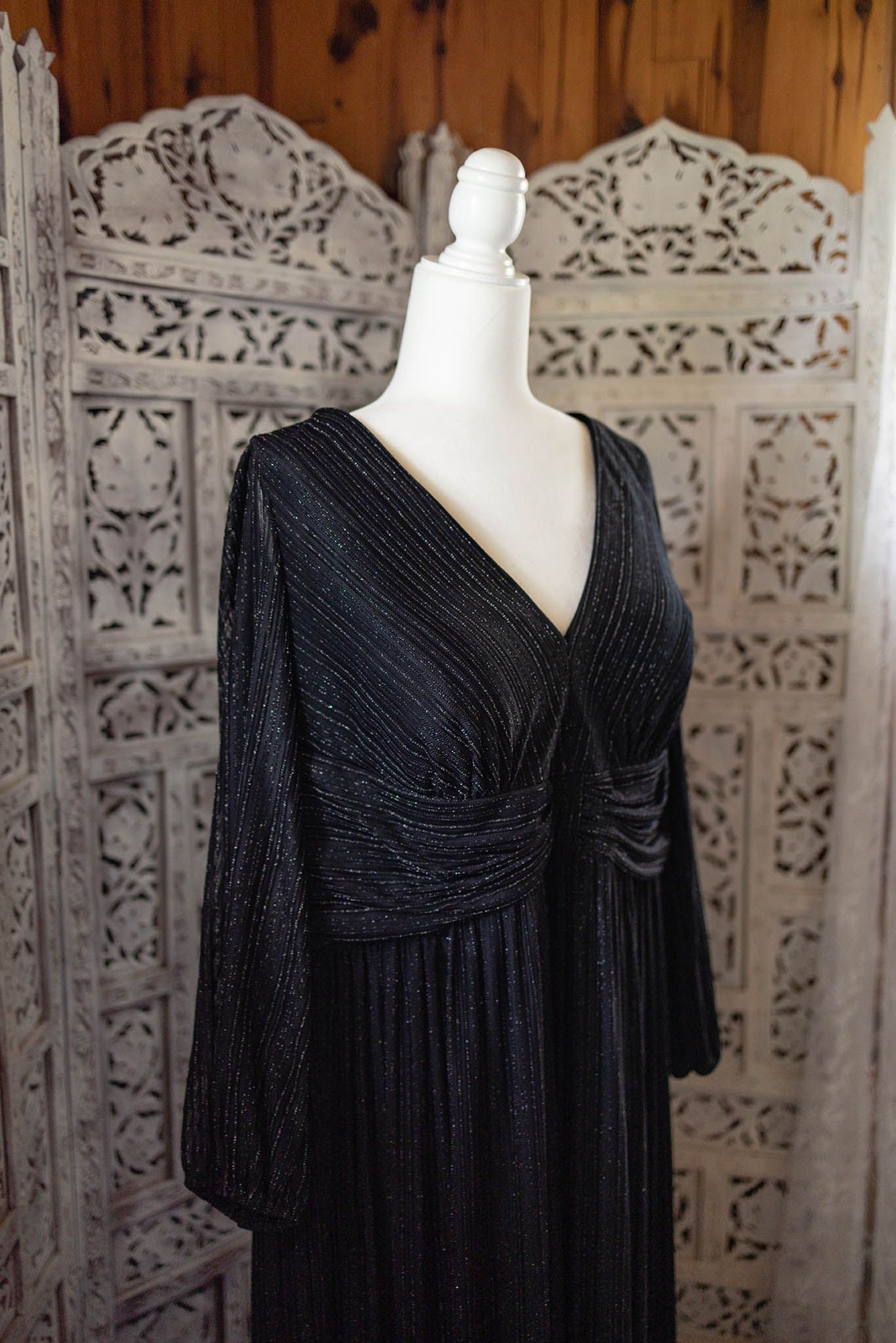 Elegant black evening gown with long sleeves and V-neck displayed on white mannequin against ornate wooden screen.