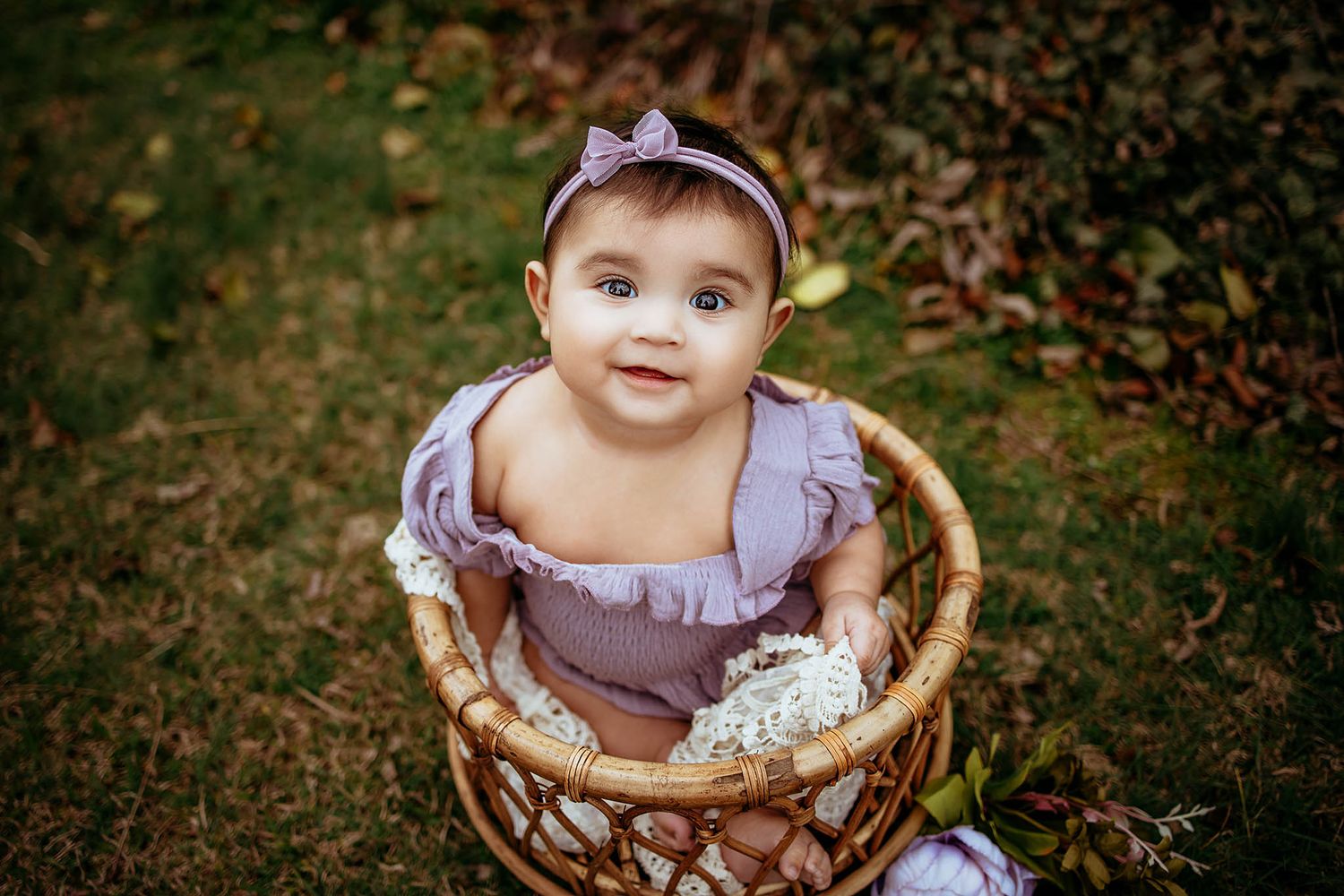 Baby in purple dress sitting in rustic wooden basket outdoors.