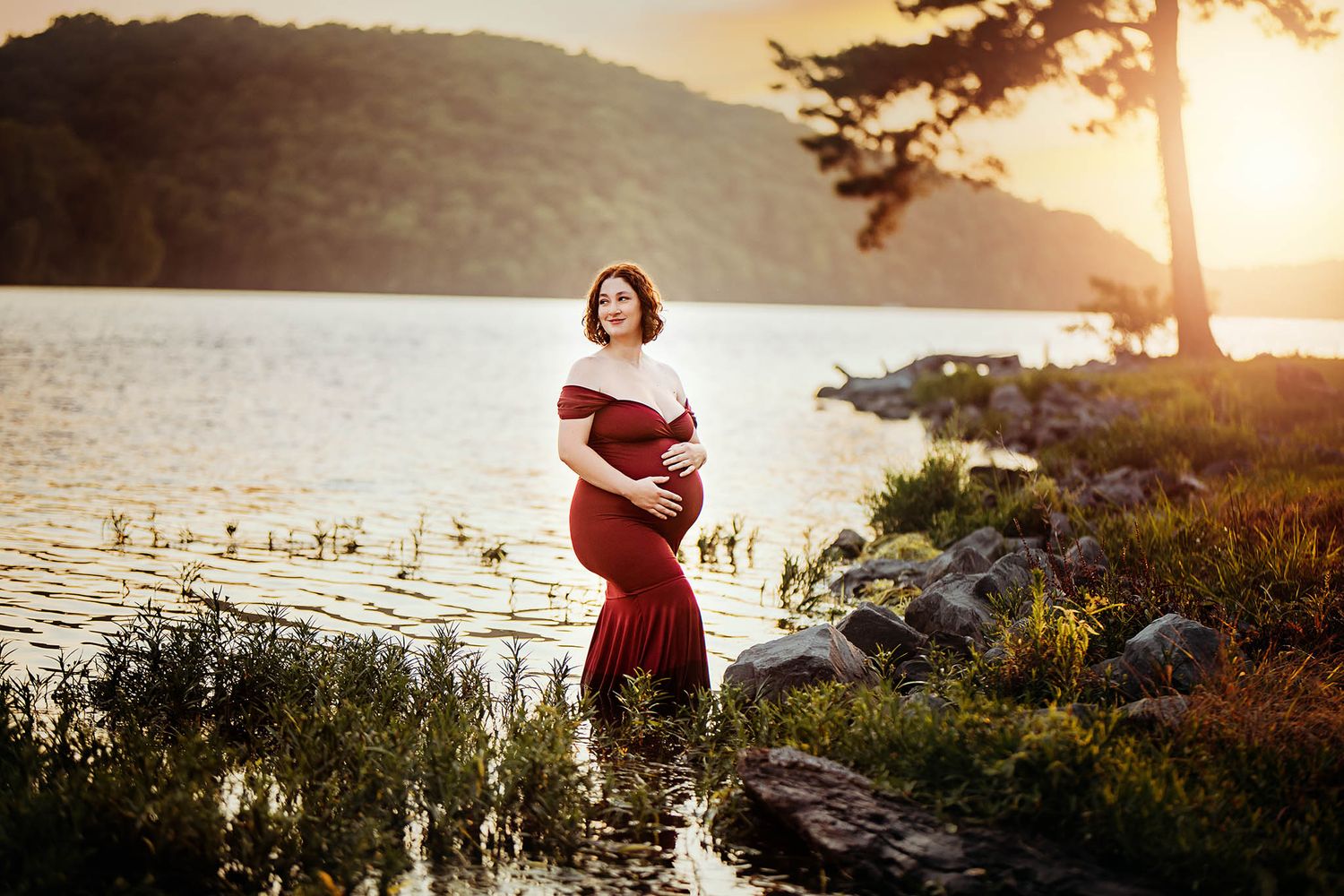 Woman in red dress poses for maternity photo by lake during golden hour.