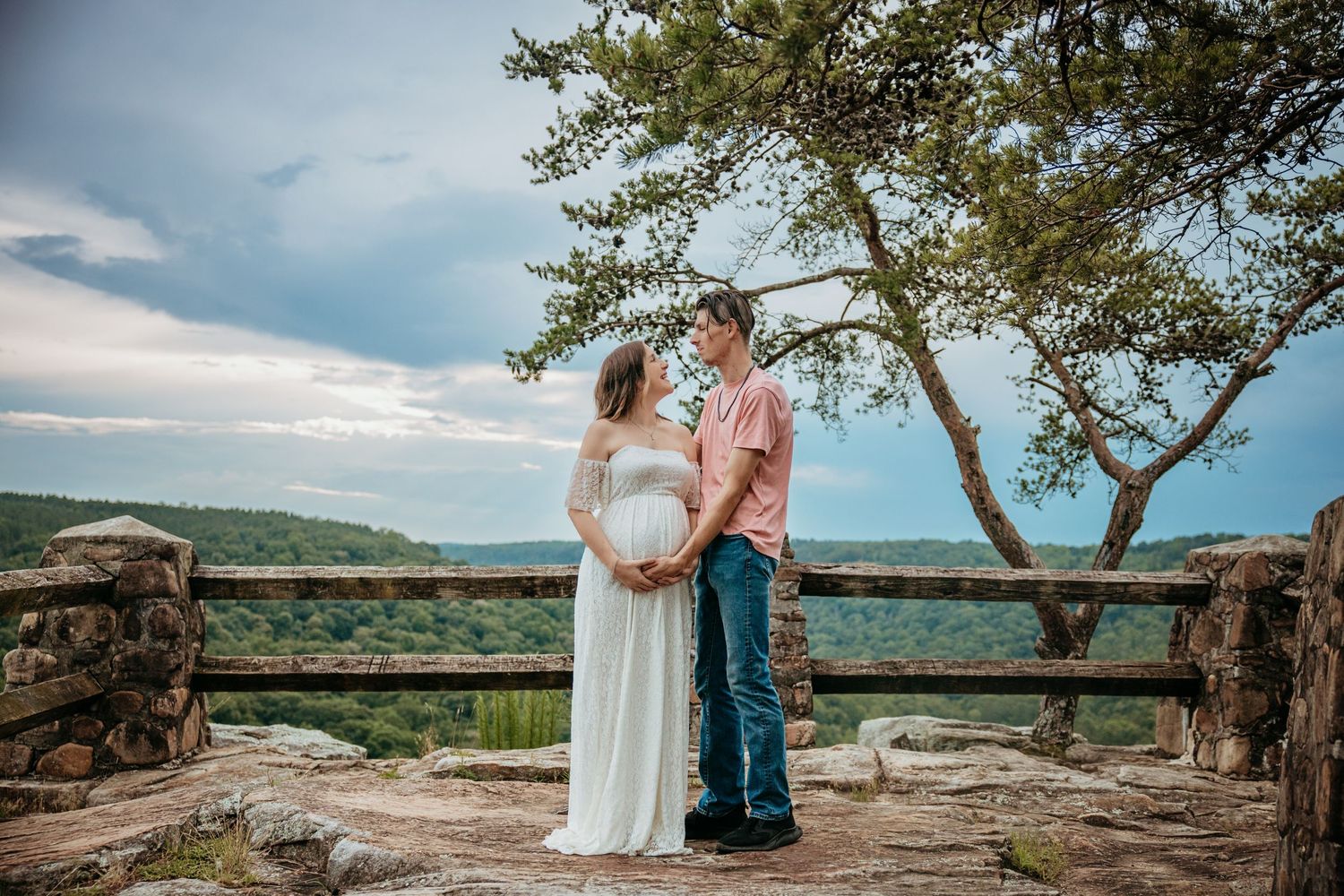 Couple sharing a tender maternity moment on a scenic overlook with lush green hills and cloudy sky backdrop.