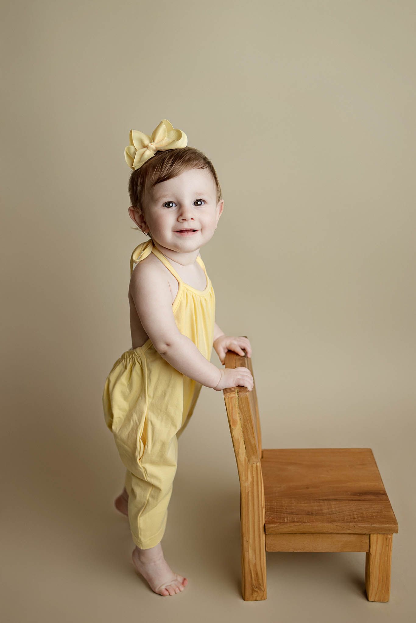 Baby in yellow romper practicing standing while holding wooden step stool.