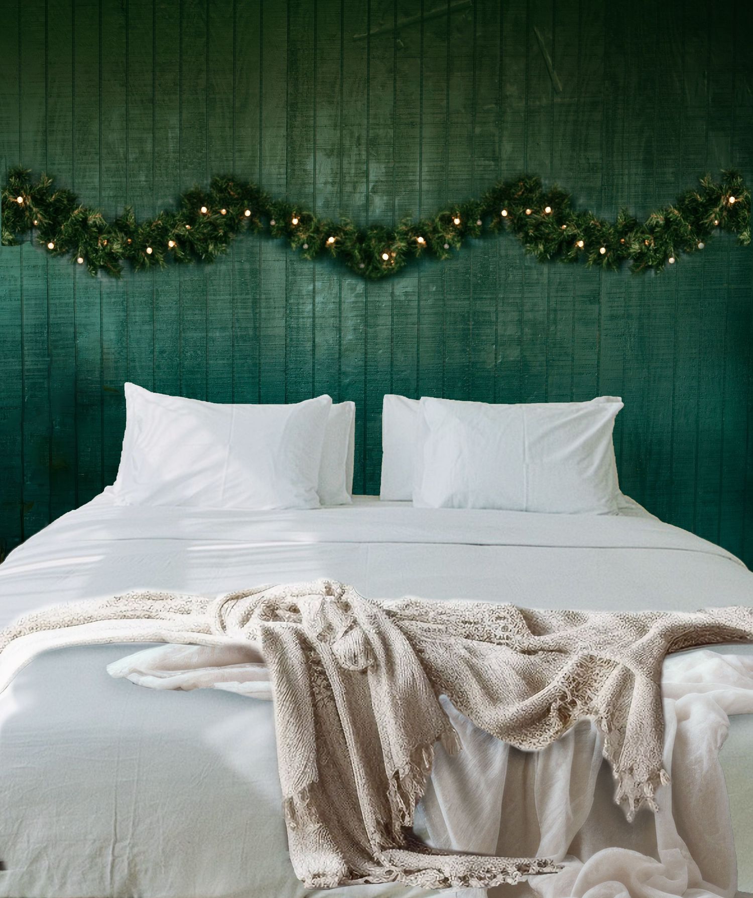 A cozy bedroom with white bedding, a beige knit throw, and a lit Christmas garland draped on a dark green shiplap wall.