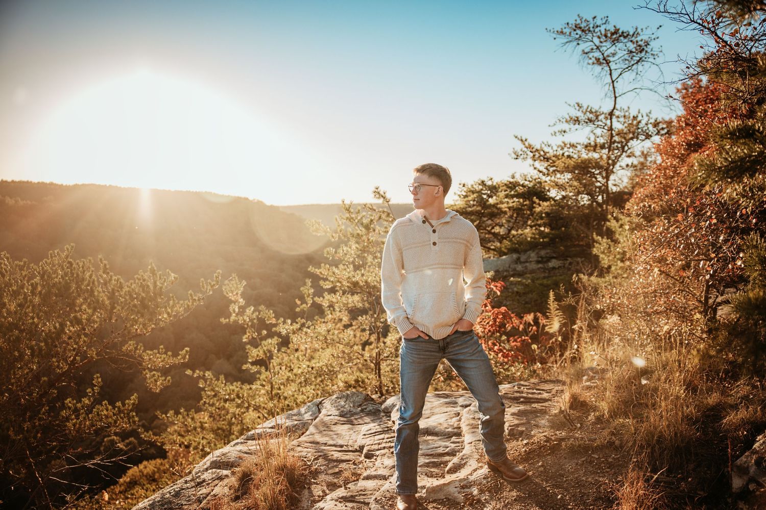 Man standing on rocky overlook at golden hour with autumn trees and glowing sunlight in a scenic mountain landscape.