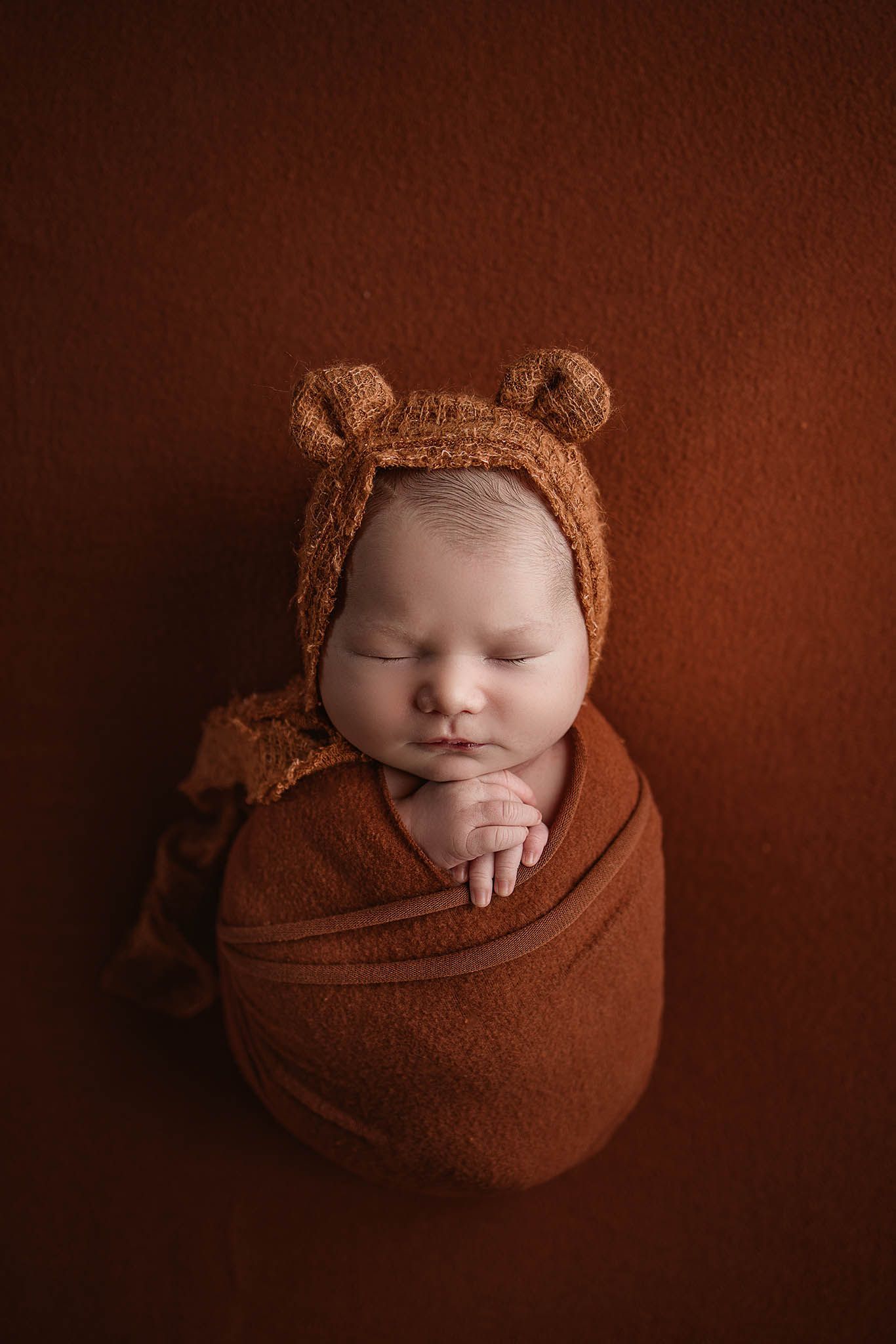 Baby wrapped in brown fabric wearing teddy bear bonnet against chocolate brown background.