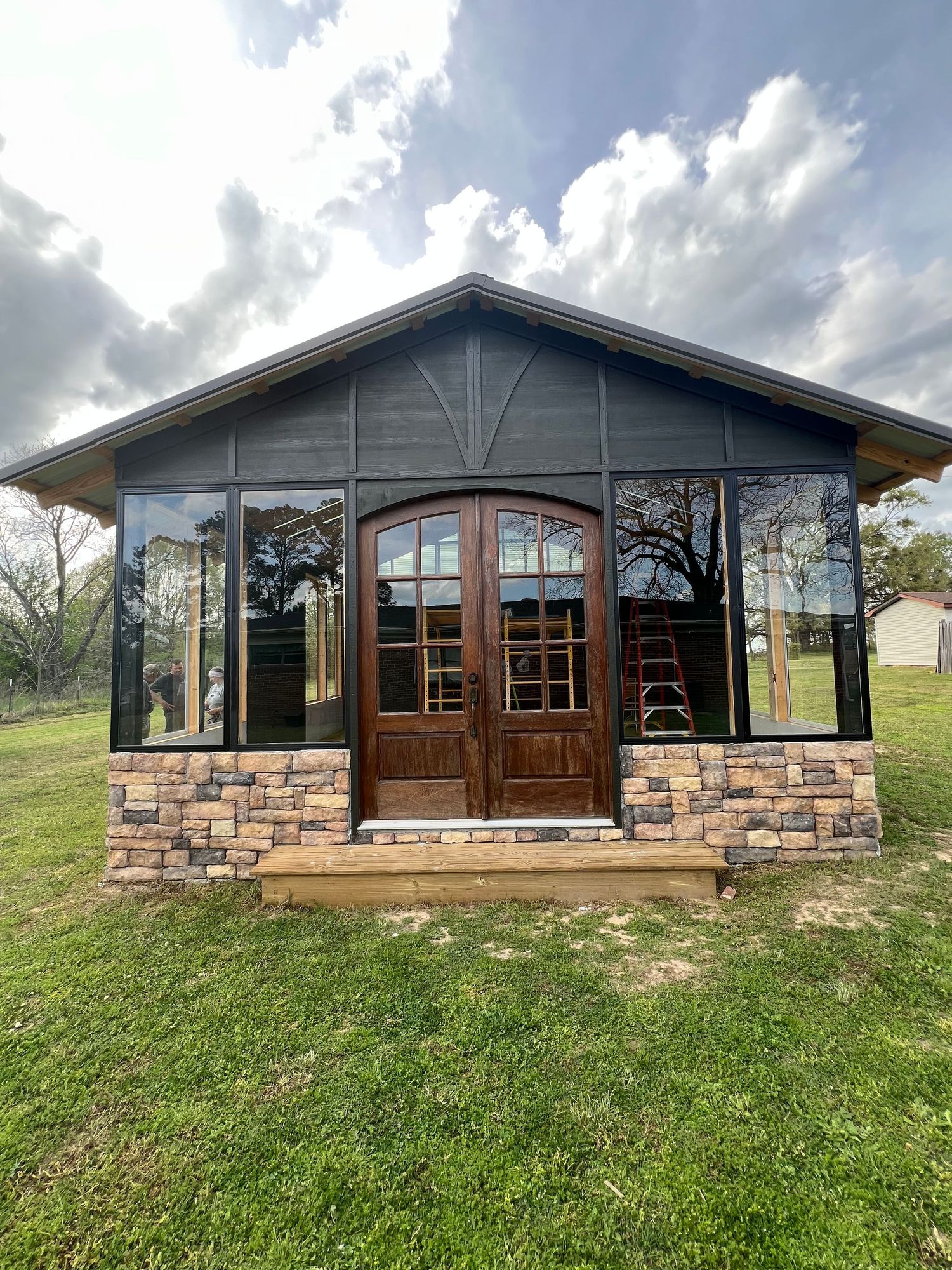 Panoramic view of a rustic wooden cabin with stone foundation nestled on a grassy hillside under dramatic cloudy skies.