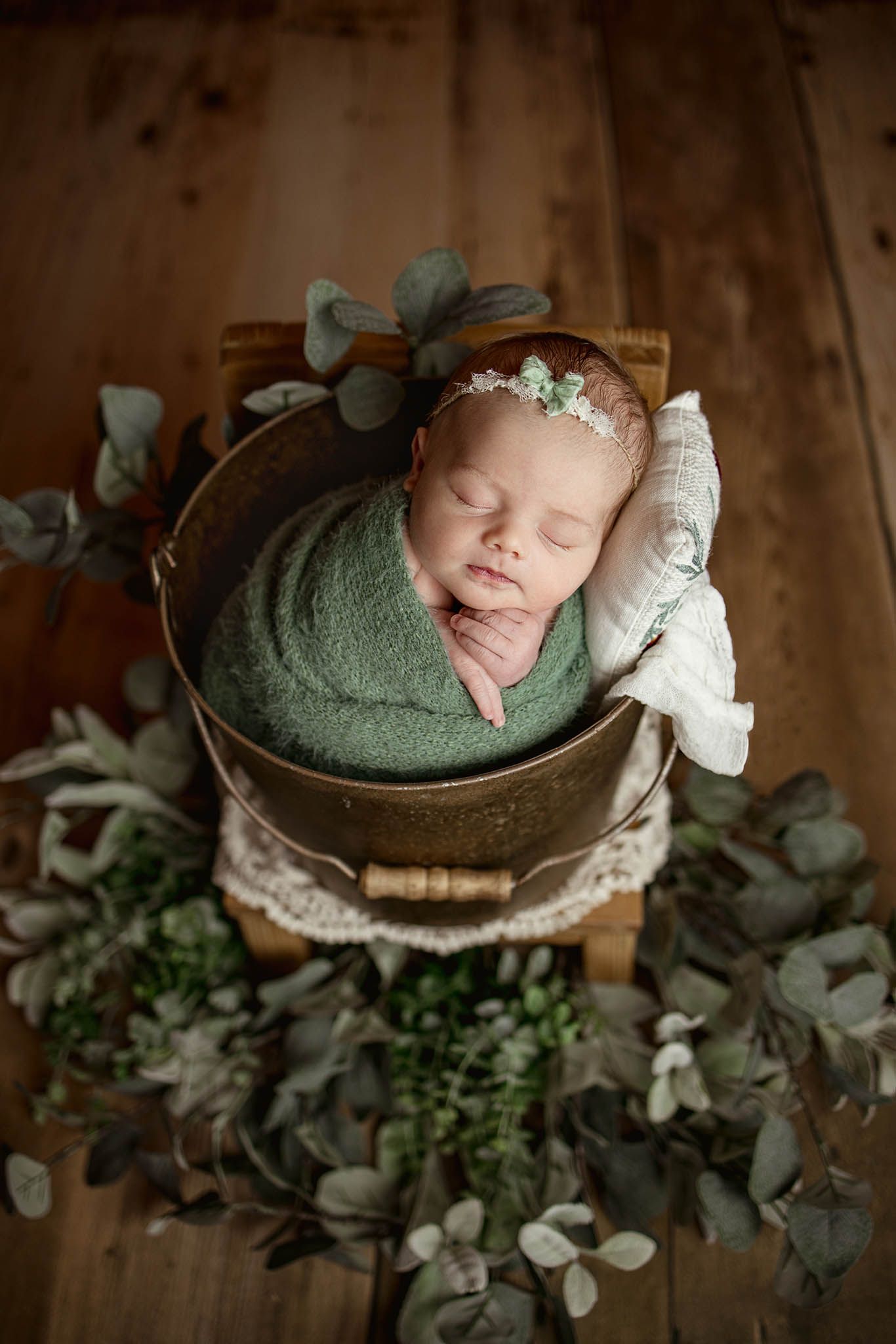 Sleeping baby wrapped in green swaddle nestled in wooden bucket with eucalyptus leaves.