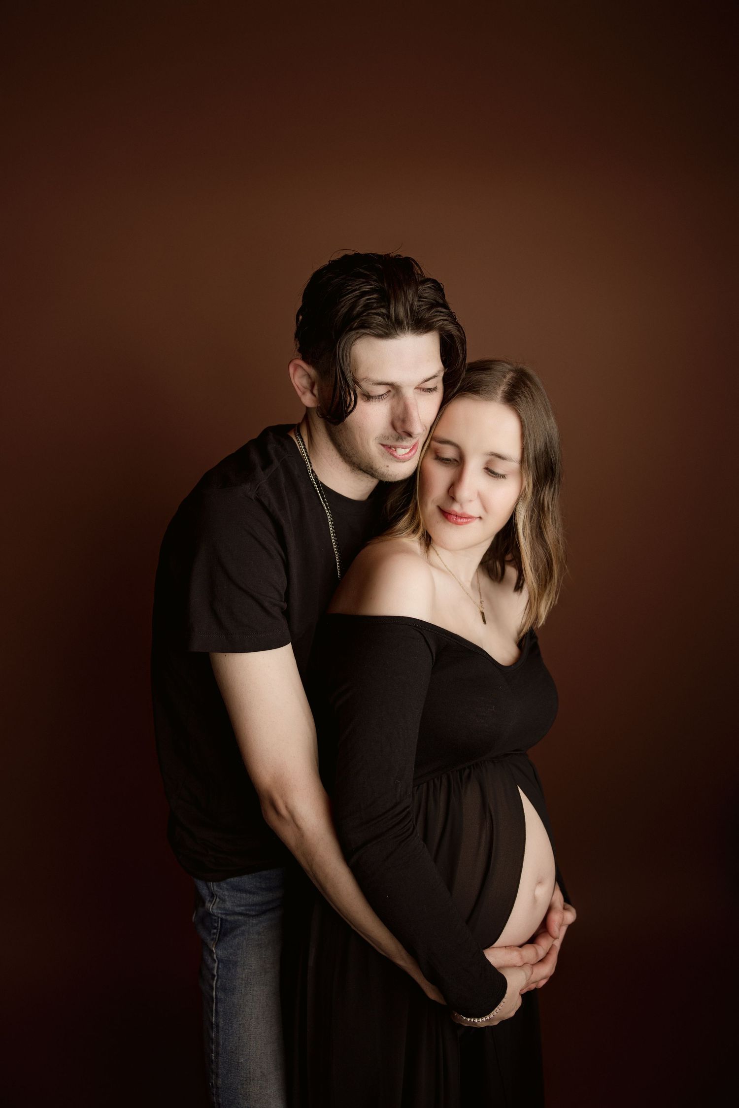 Intimate maternity portrait of couple in black against dark brown background.