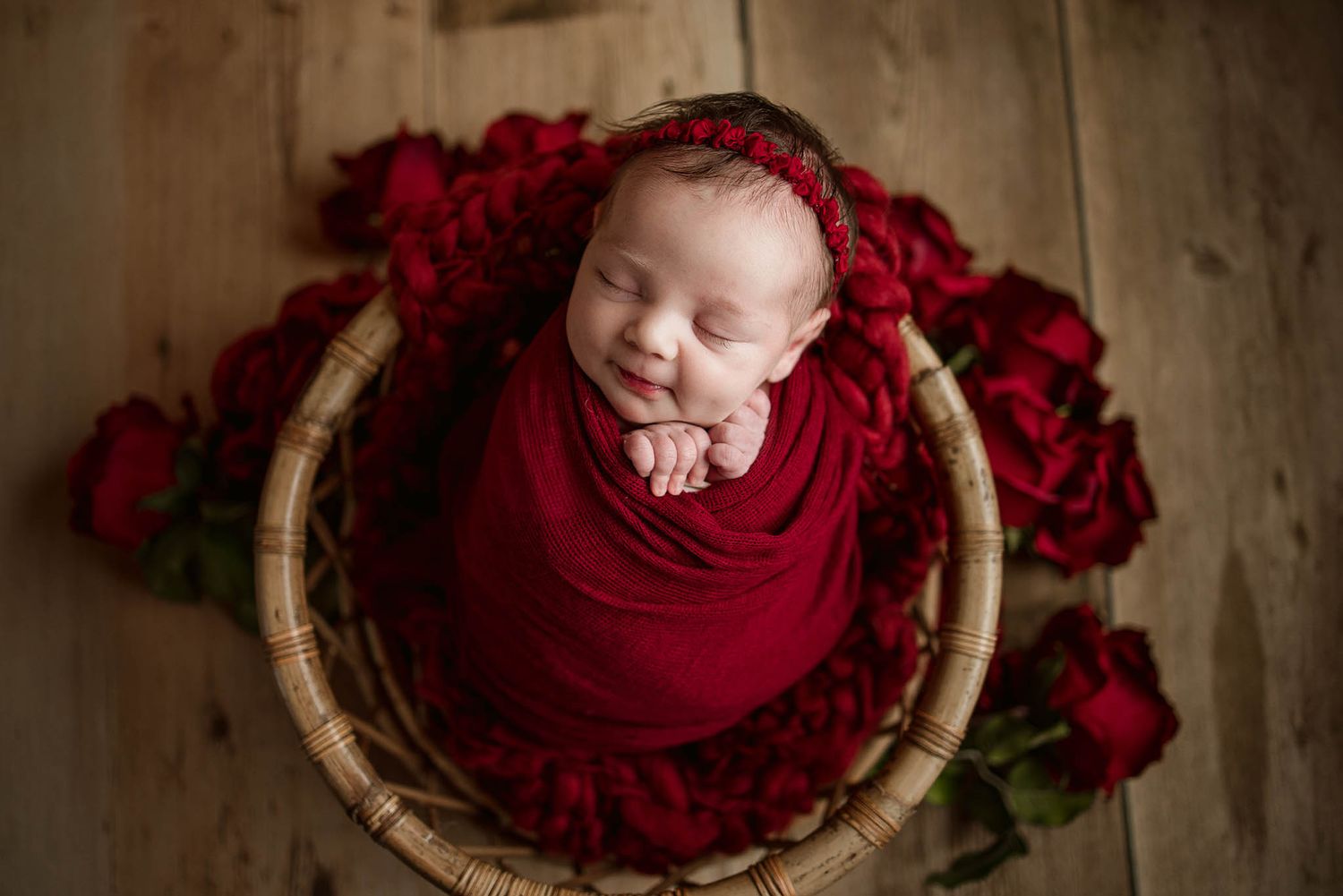 Sleeping newborn wrapped in deep red fabric surrounded by red roses in woven basket.