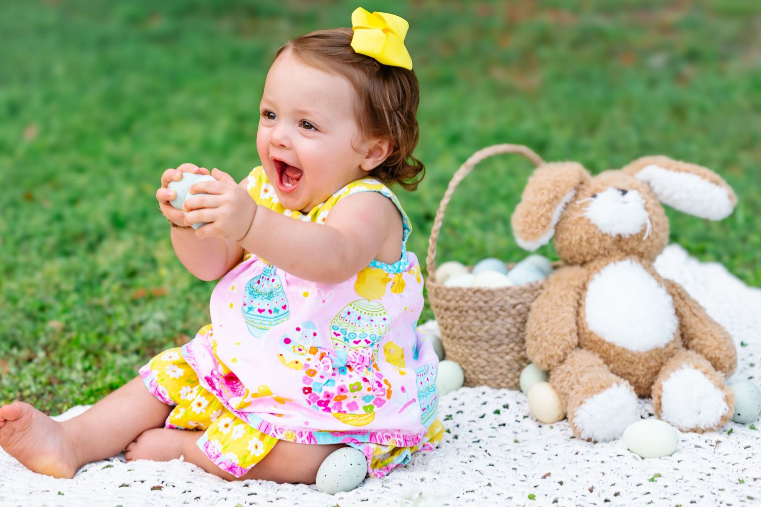 Baby plays on white blanket with stuffed animals and toys in garden setting.