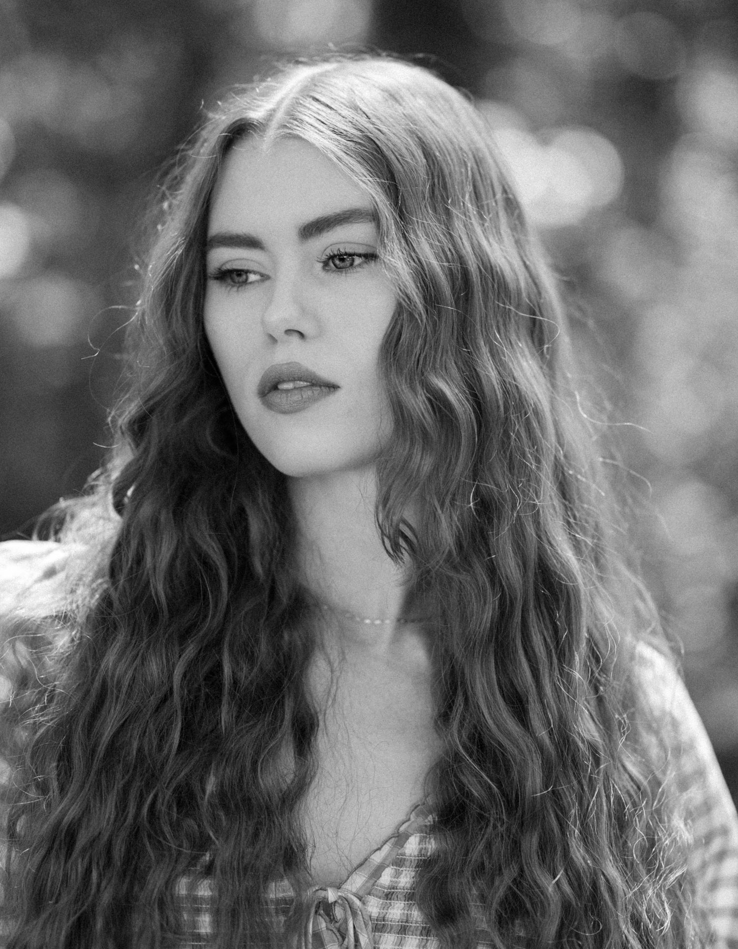 Black and white portrait of a young woman with long wavy hair, wearing a plaid top outdoors.