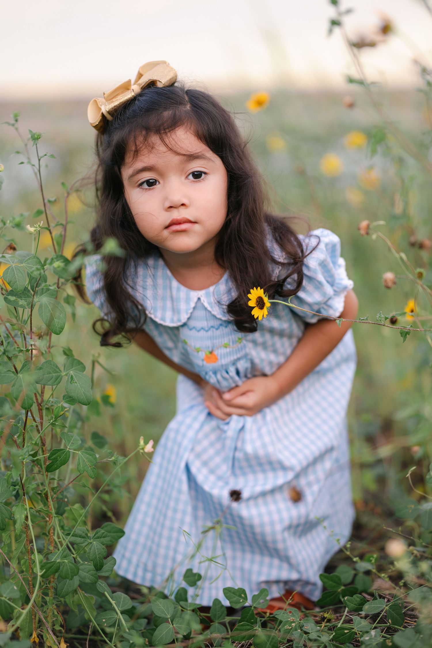 A child in a gingham dress bends down among yellow wildflowers in a field.