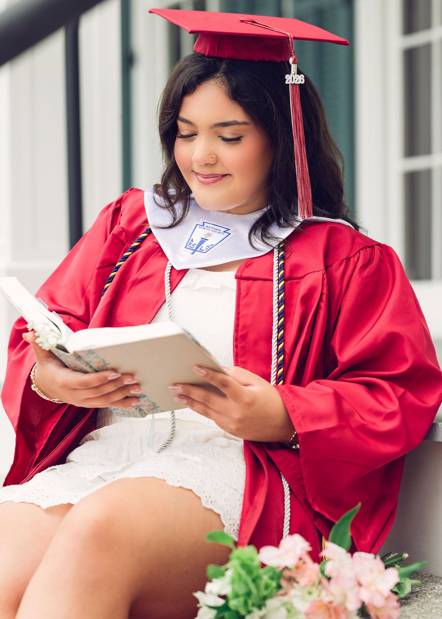Graduate in red cap and gown sits outdoors holding a book while surrounded by pink flowers and greenery.