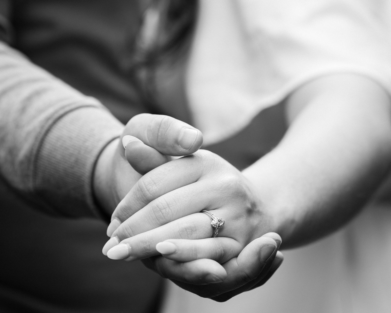 Black and white close-up of two people holding hands, one wearing a diamond engagement ring.
