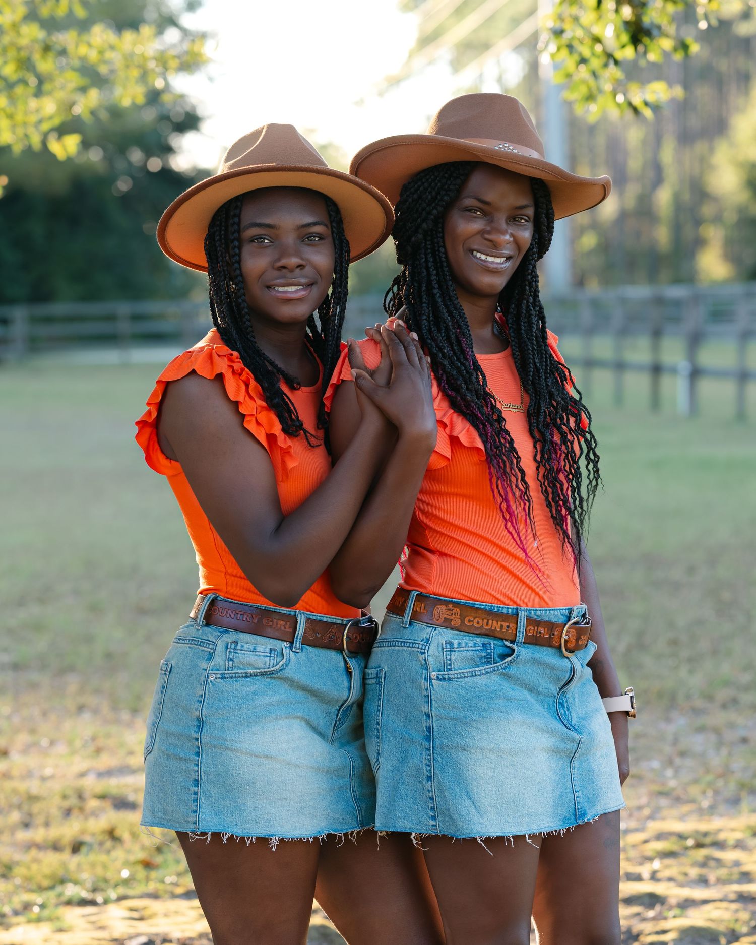 Two friends in matching orange tops and denim shorts pose together wearing brown cowboy hats.