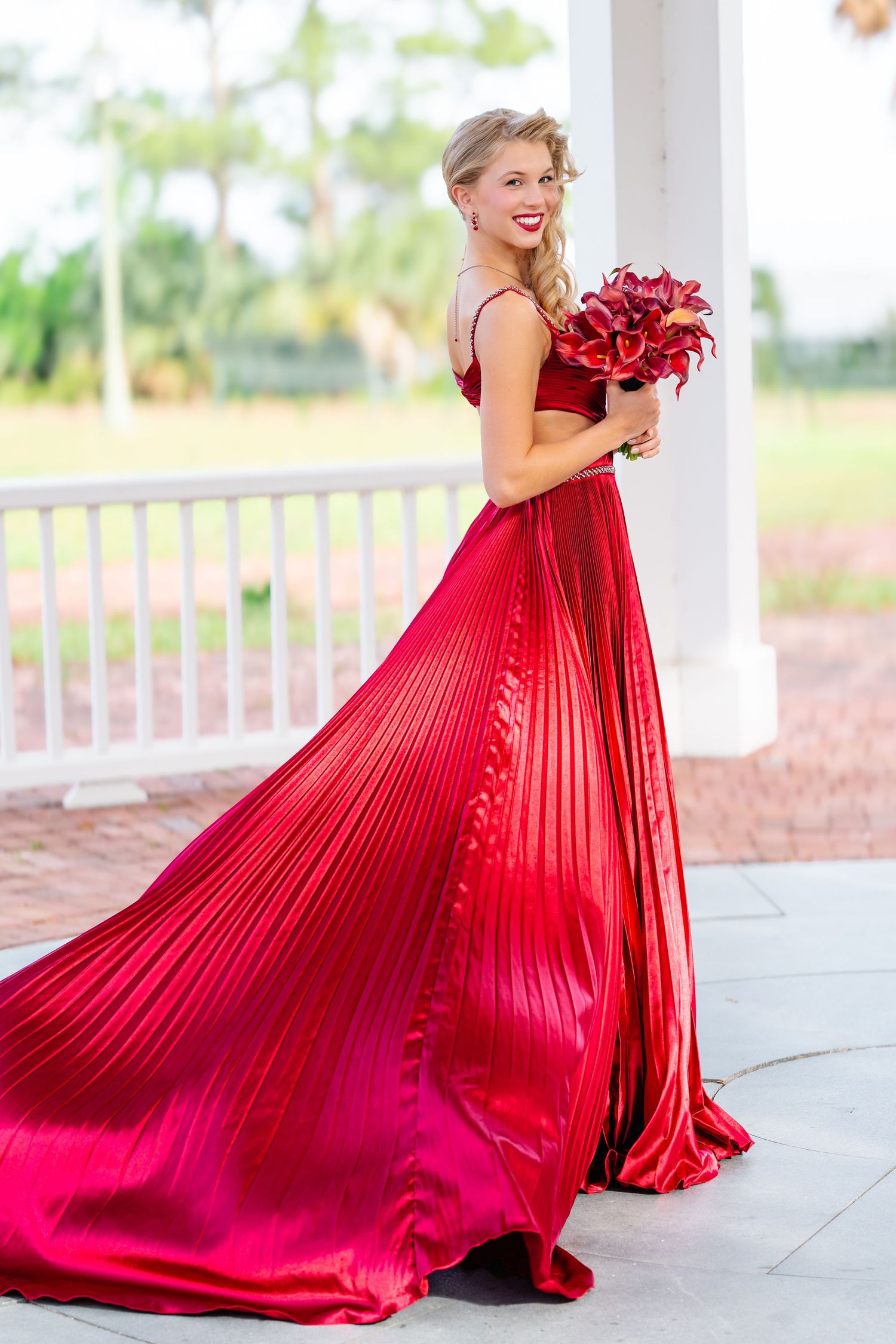 Flowing red pleated gown with matching bouquet photographed on an elegant white porch.