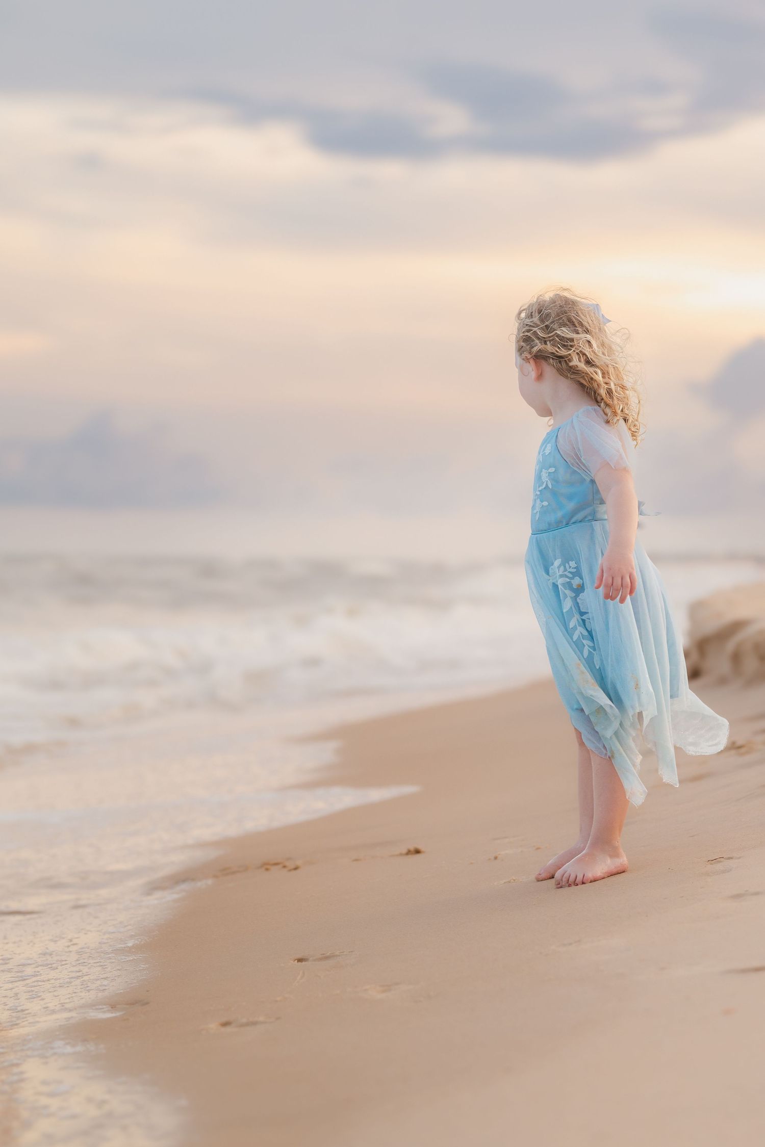 A person in a flowing light blue dress walks barefoot along a serene beach at sunset.