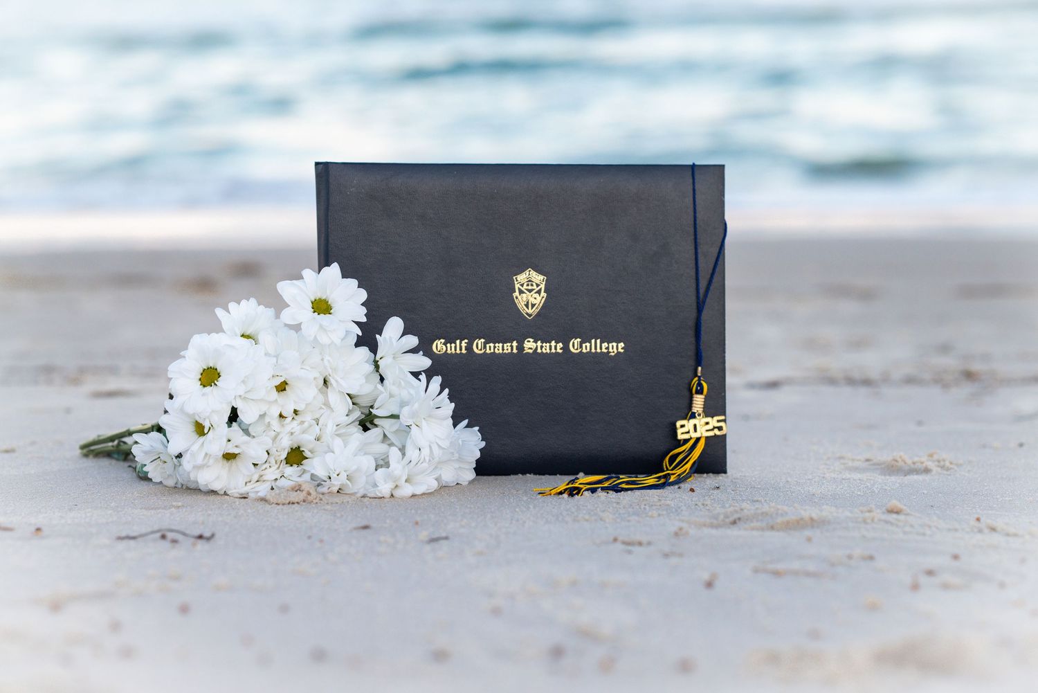 Black graduation cap with gold tassel and white flowers on sandy beach.