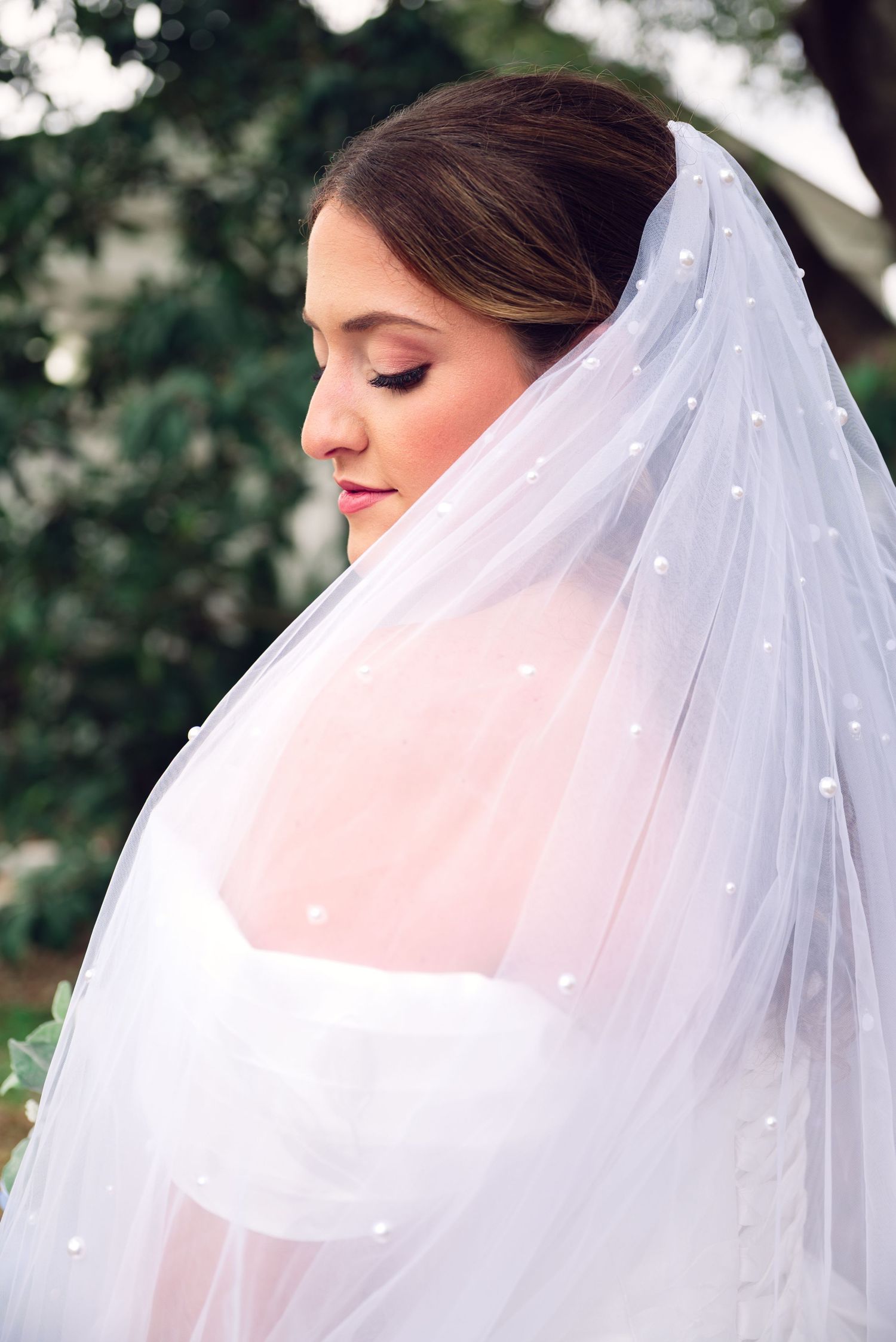 Bride in white wedding dress and pearl-adorned veil with eyes closed, elegant updo hairstyle and soft makeup outdoors.