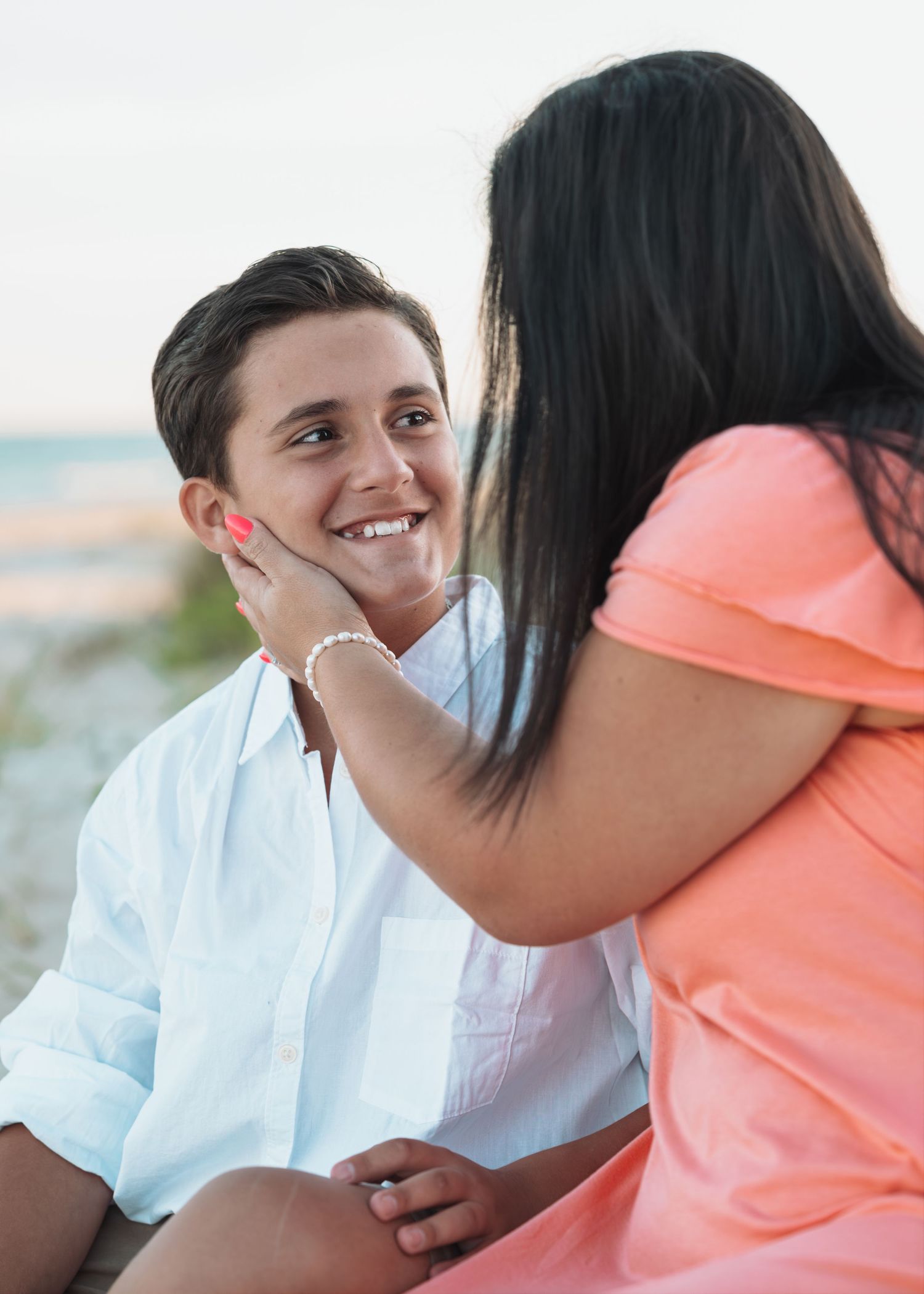A smiling young man in a white shirt gazes at a woman in a coral dress touching his face on a beach.