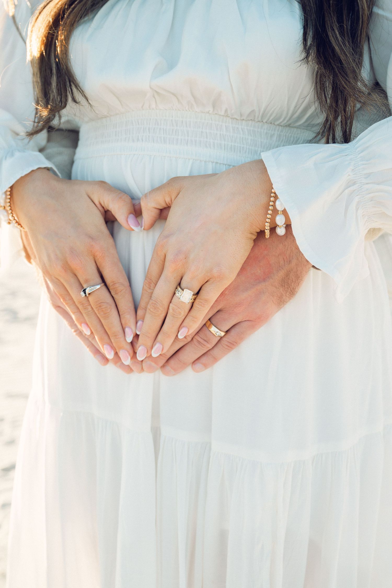 Close-up of hands wearing delicate gold rings and pearl jewelry against flowing white fabric.