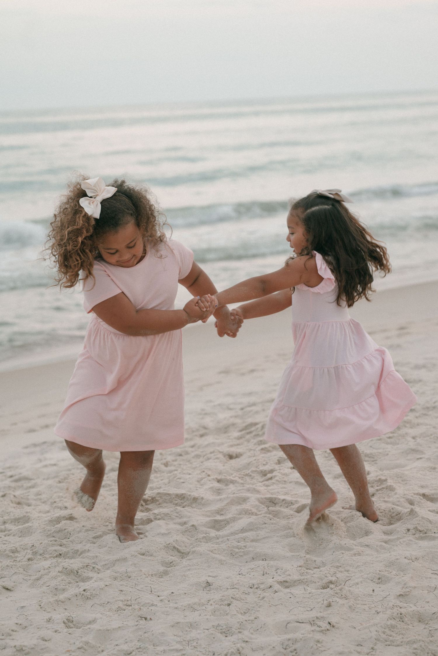 Two young girls in pink dresses hold hands and spin together on a sandy beach near the ocean.