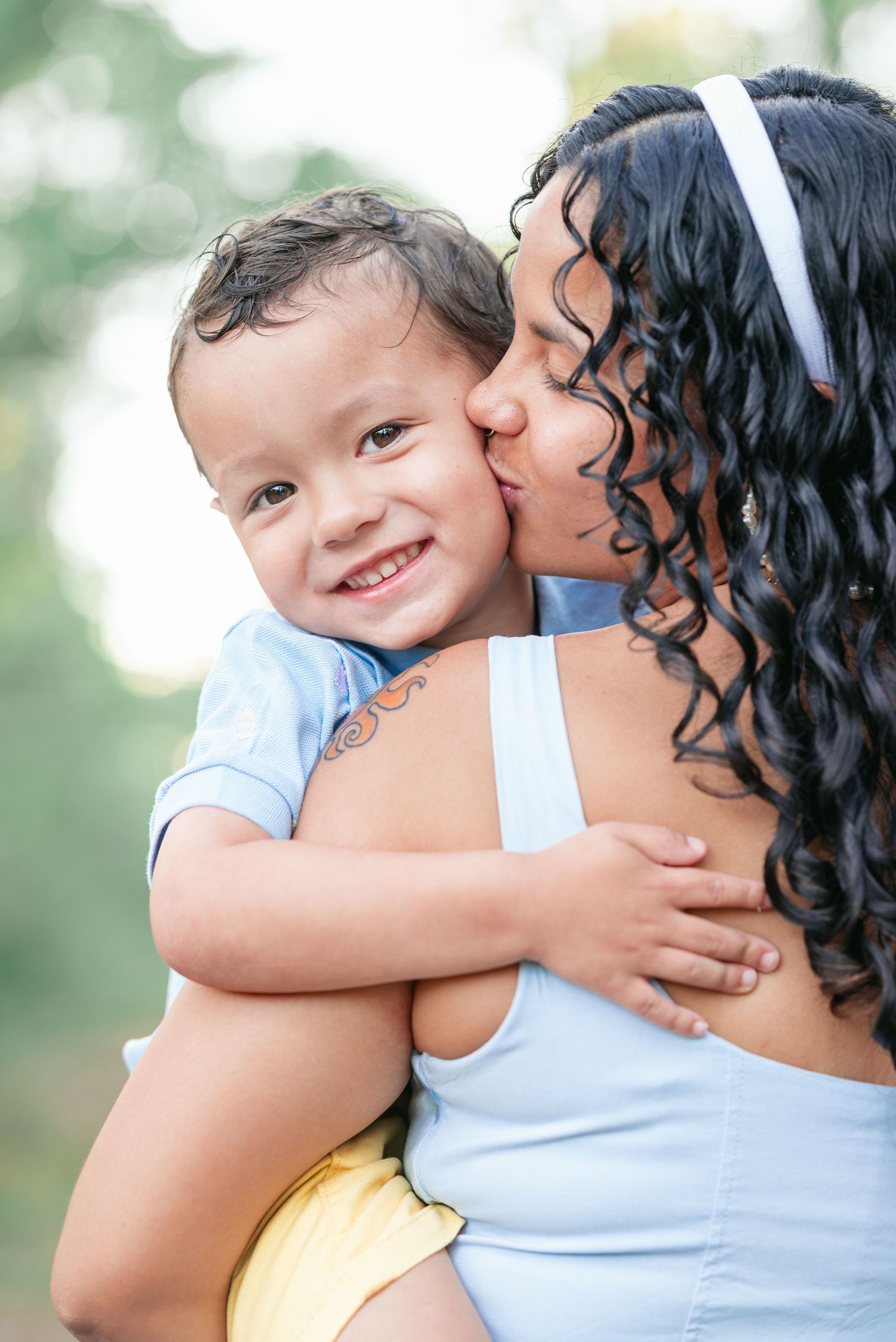 A curly-haired woman in a blue top kisses a smiling toddler boy outdoors in a warm, tender moment.