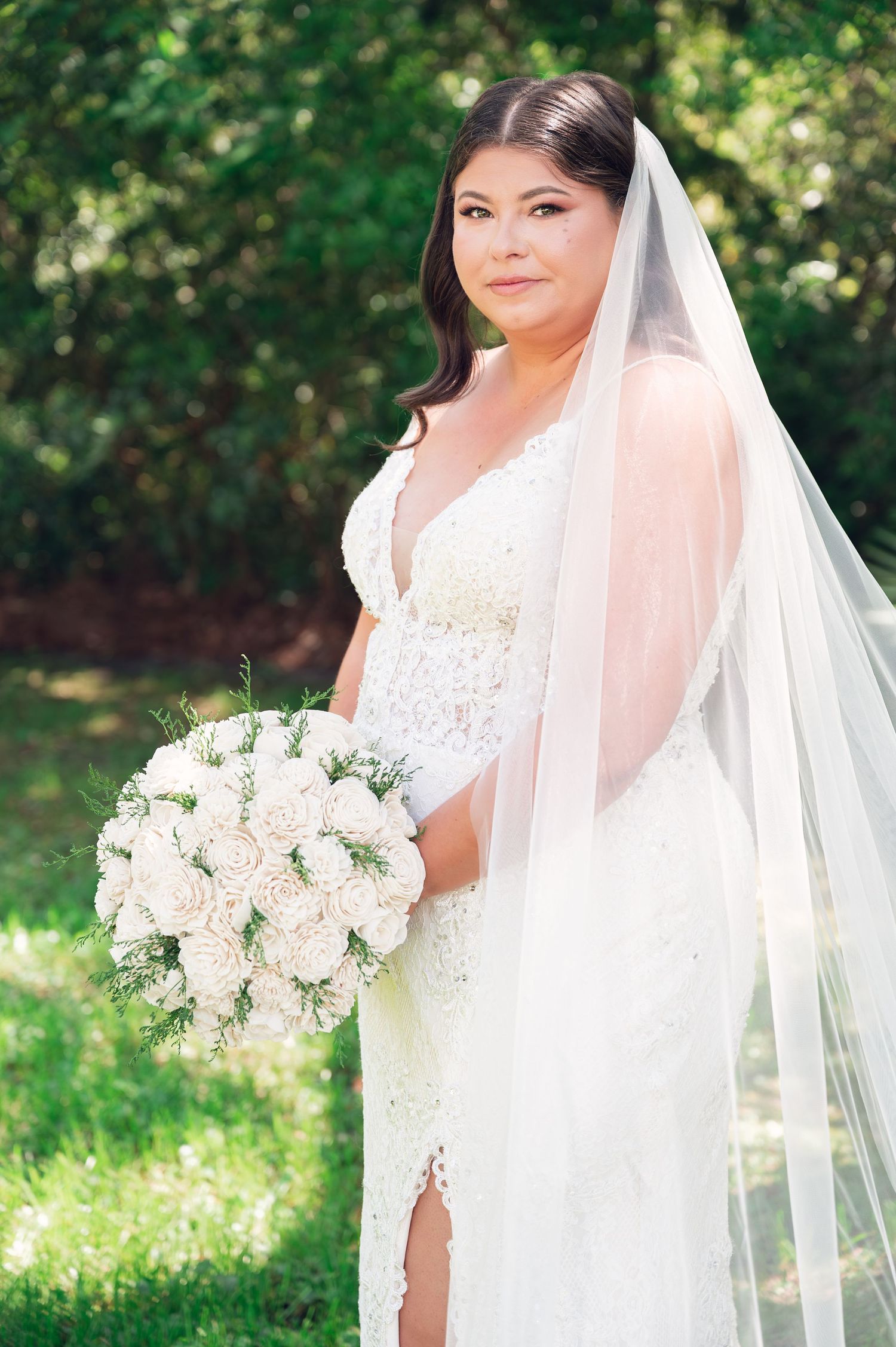 Bride in white lace gown and veil holding white rose bouquet, posing outdoors with green trees background.