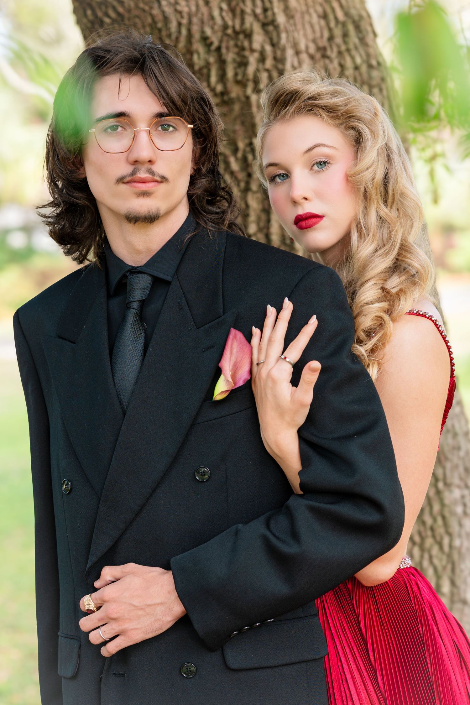 Stylish couple in black suit and red dress pose playfully against a tree backdrop.