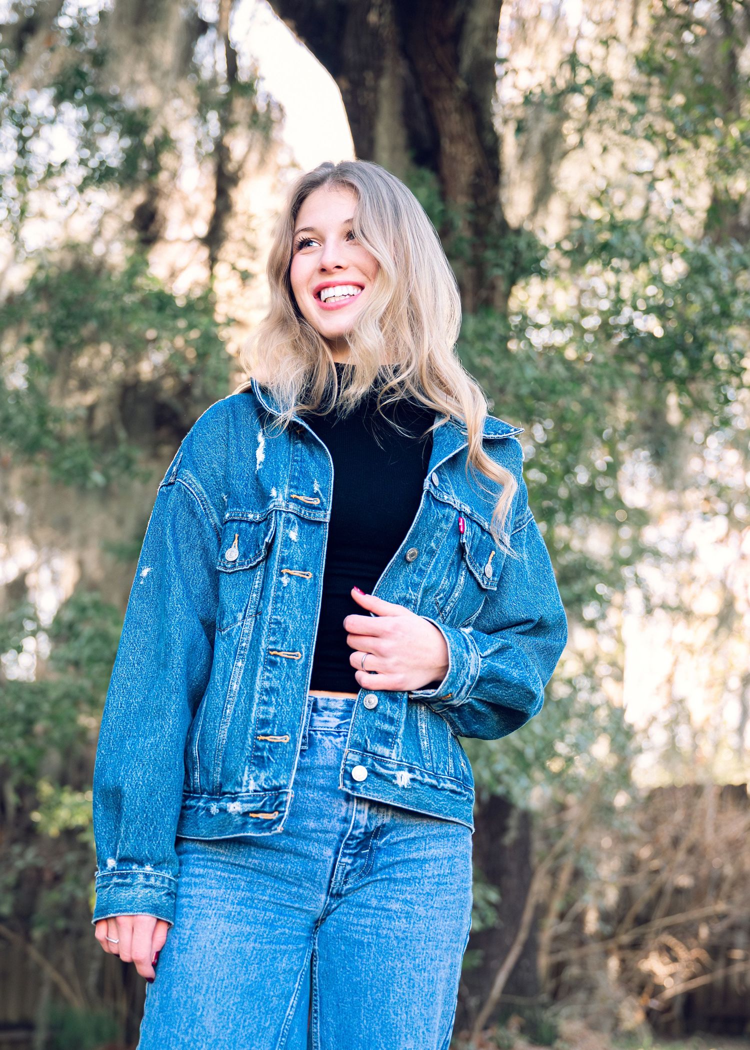 Casual outdoor fashion photoshoot featuring denim jacket and black top against natural foliage background.