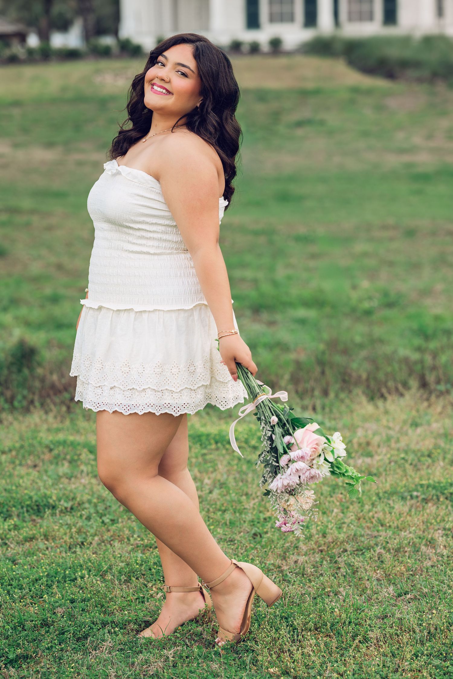 A person in a white summer dress poses playfully in a grassy field while holding a pink floral bouquet.