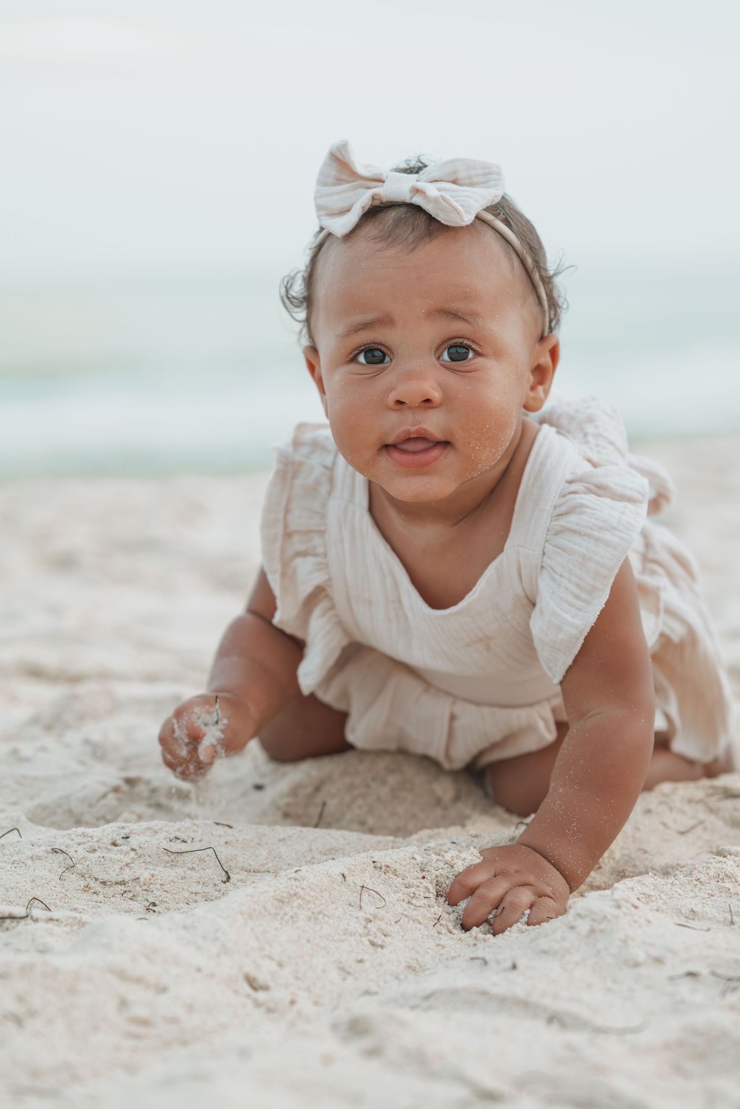 Adorable baby girl in a white ruffle outfit and bow headband crawling on sandy beach shore.