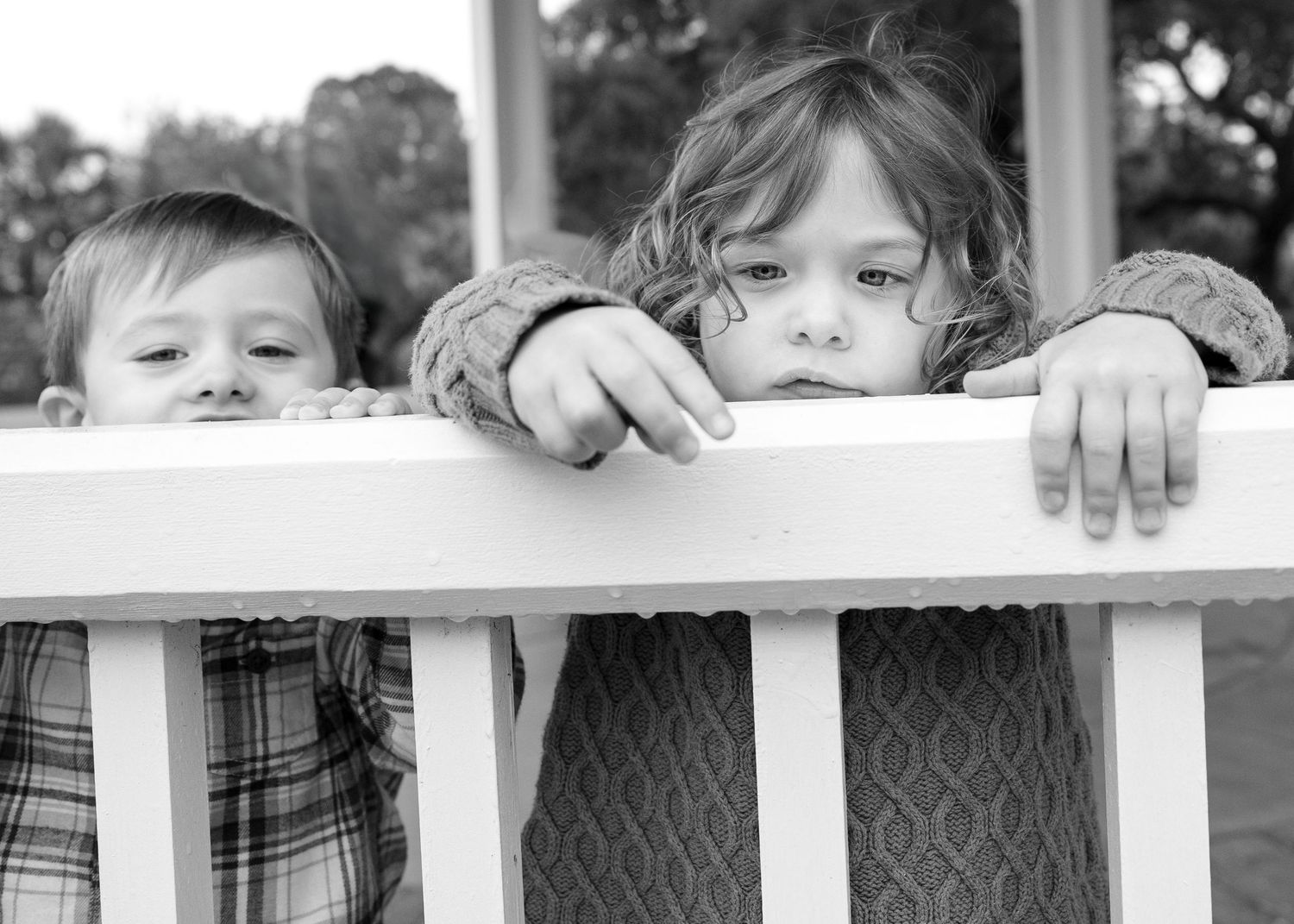 Young children peek playfully through white fence rails in black and white photograph.