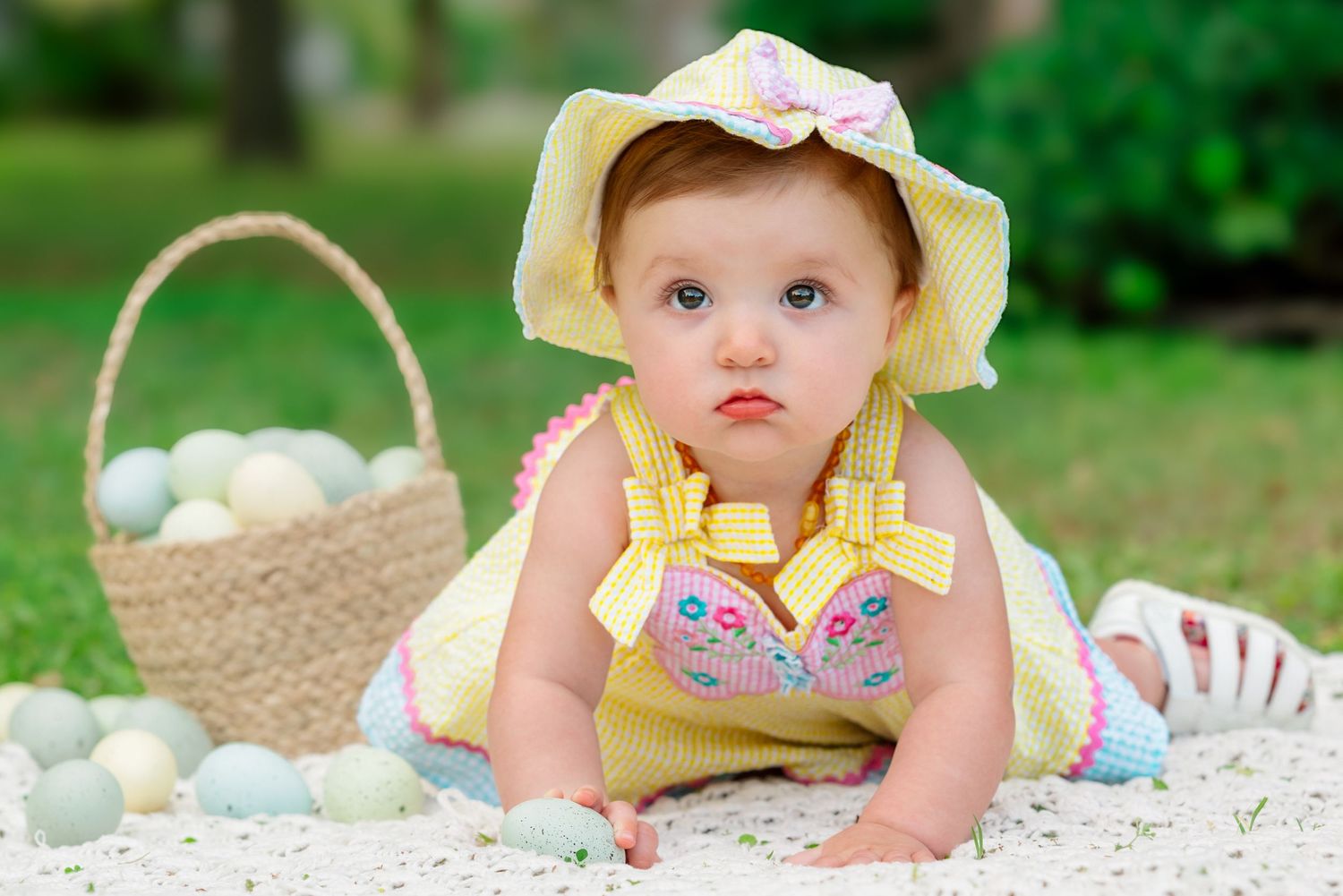 Baby in yellow sunhat and floral dress crawls on white blanket with basket nearby.