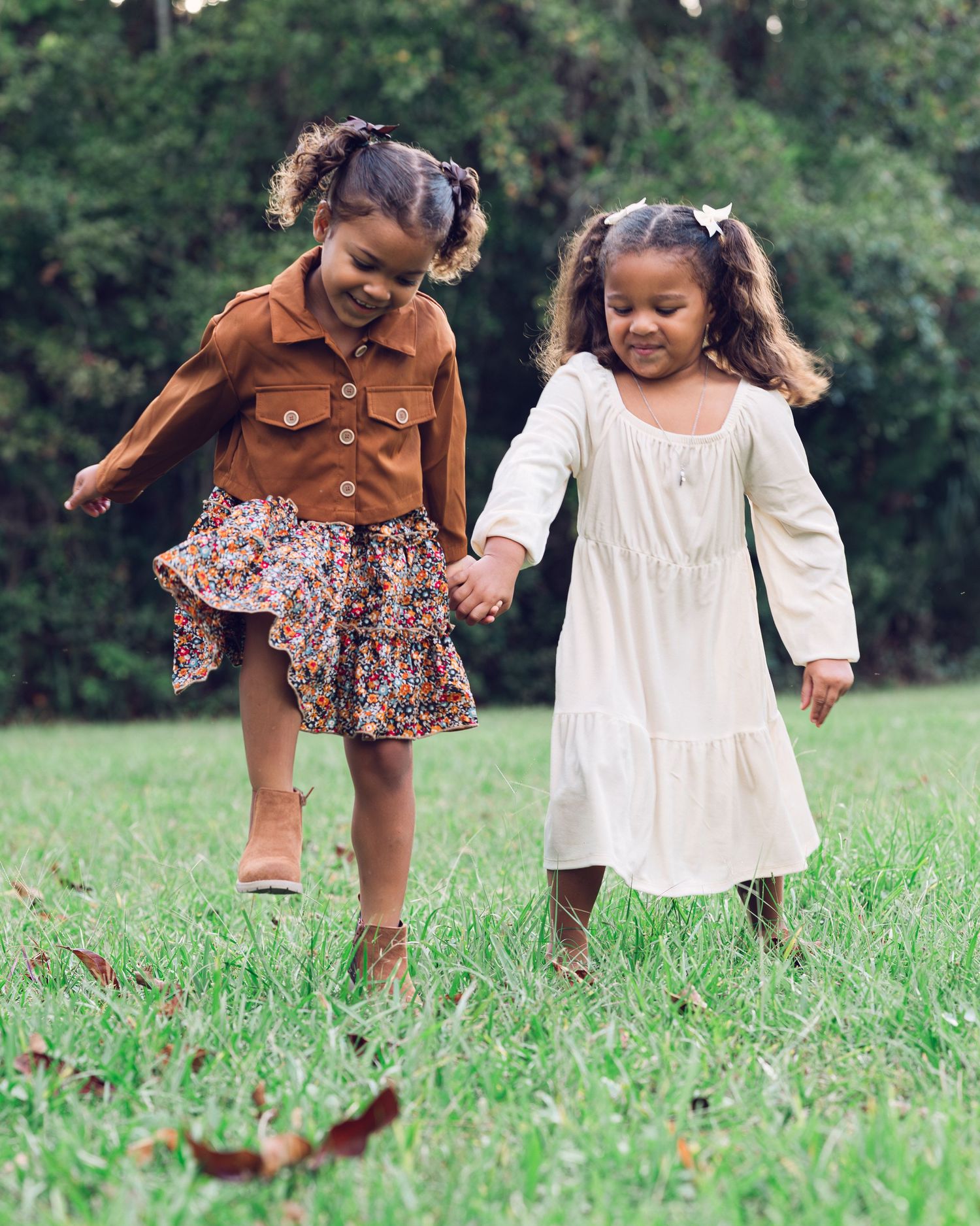 Two children in fall dresses hold hands while playing and skipping together in a grassy field.