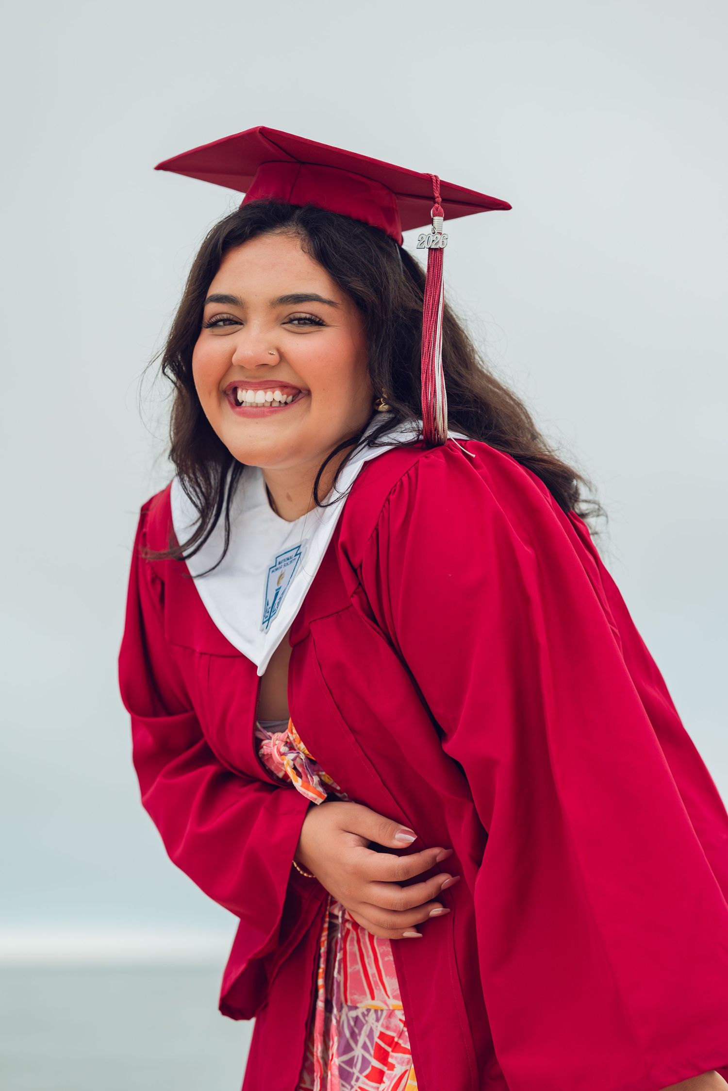 A graduate in red cap and gown smiles brightly against an overcast sky background.