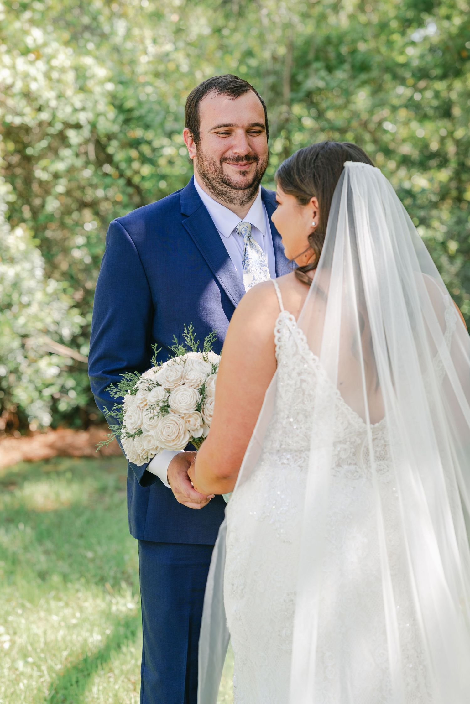 Groom in blue suit smiles at bride in lace gown holding white bouquet, outdoor wedding portrait.