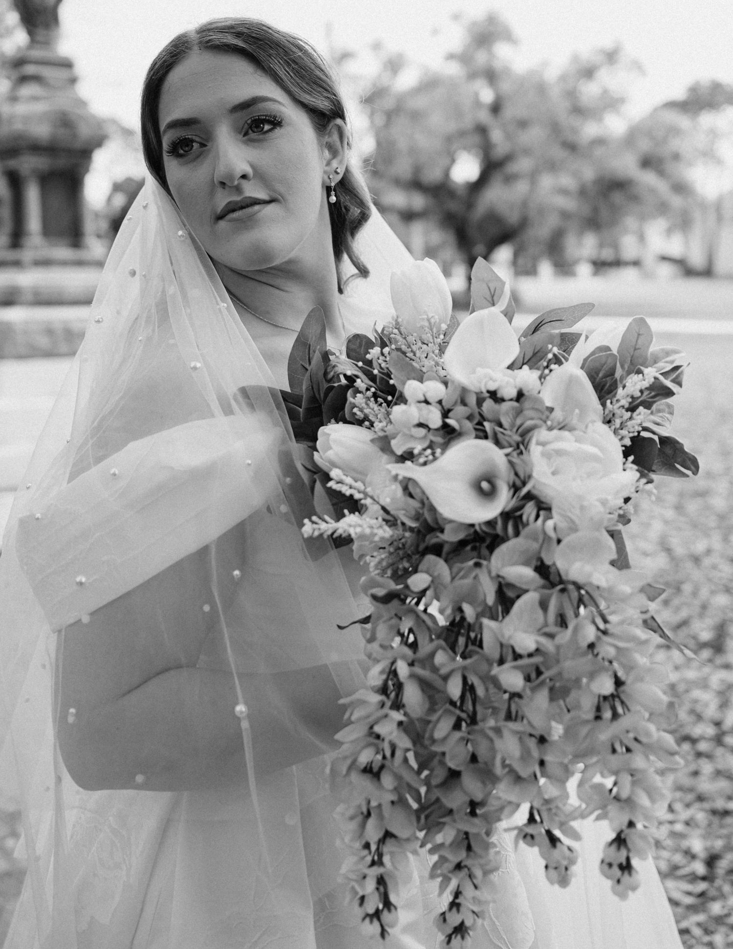 Black and white photo of a bride in a veil holding a cascading bouquet of flowers outdoors.