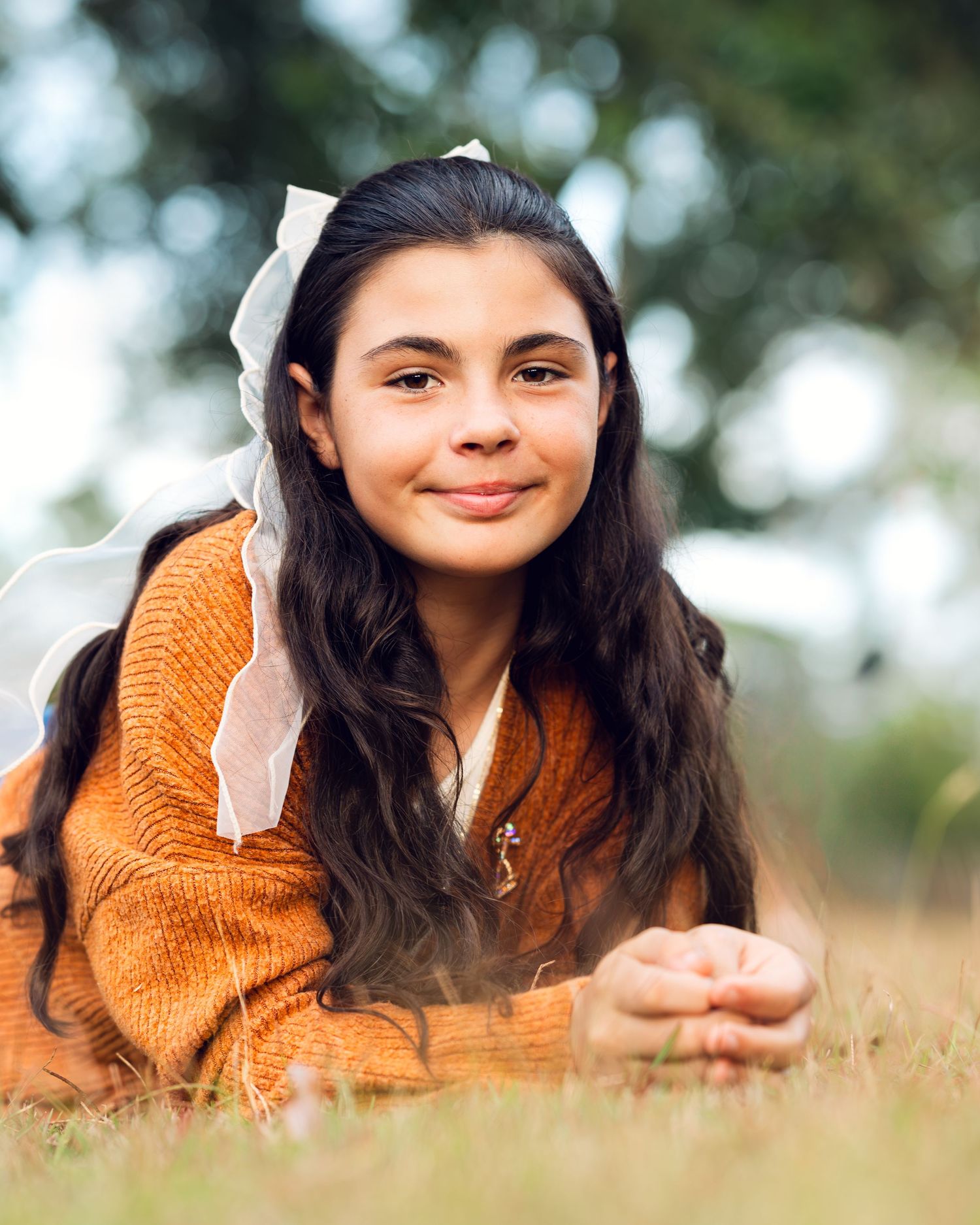 A person in an orange sweater with white bow accessory lies in autumn grass for a portrait.