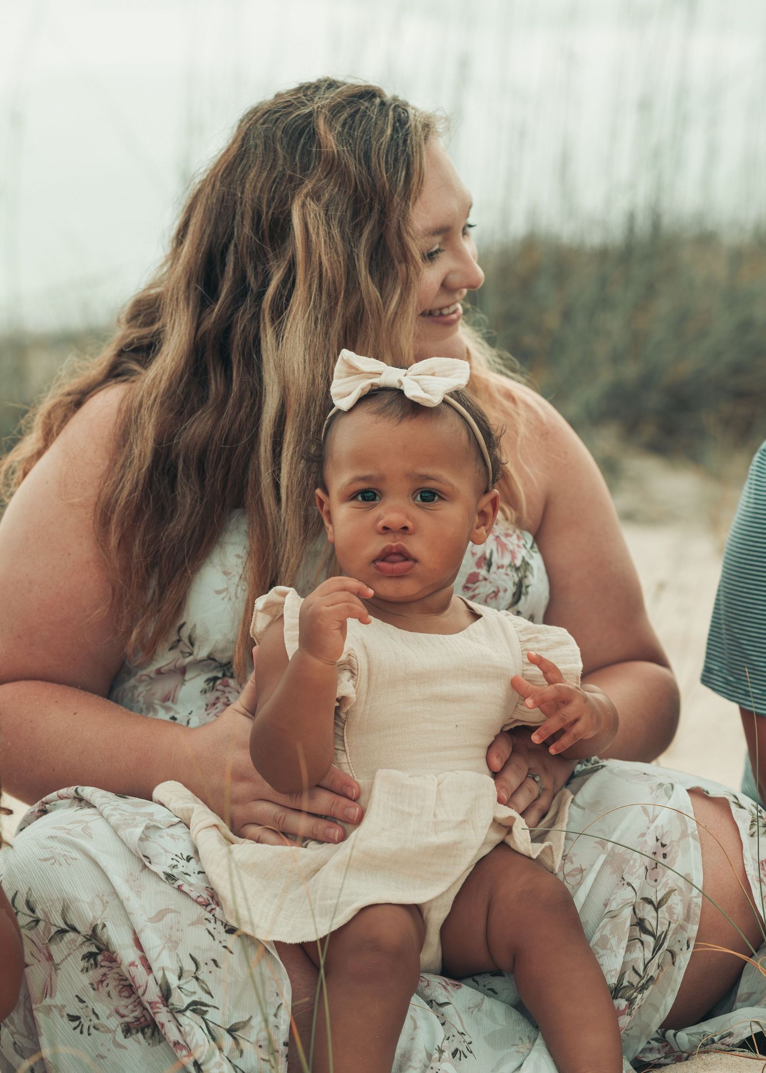A smiling woman holds a cute baby girl wearing a white bow headband and cream dress on a sandy beach.