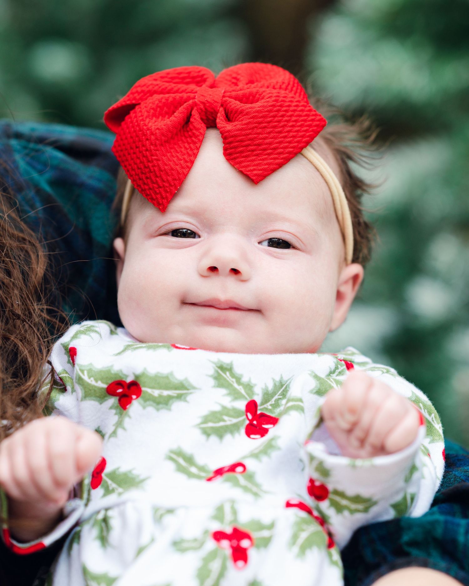 Baby wearing a festive white outfit with red bow headband against blurred green background.