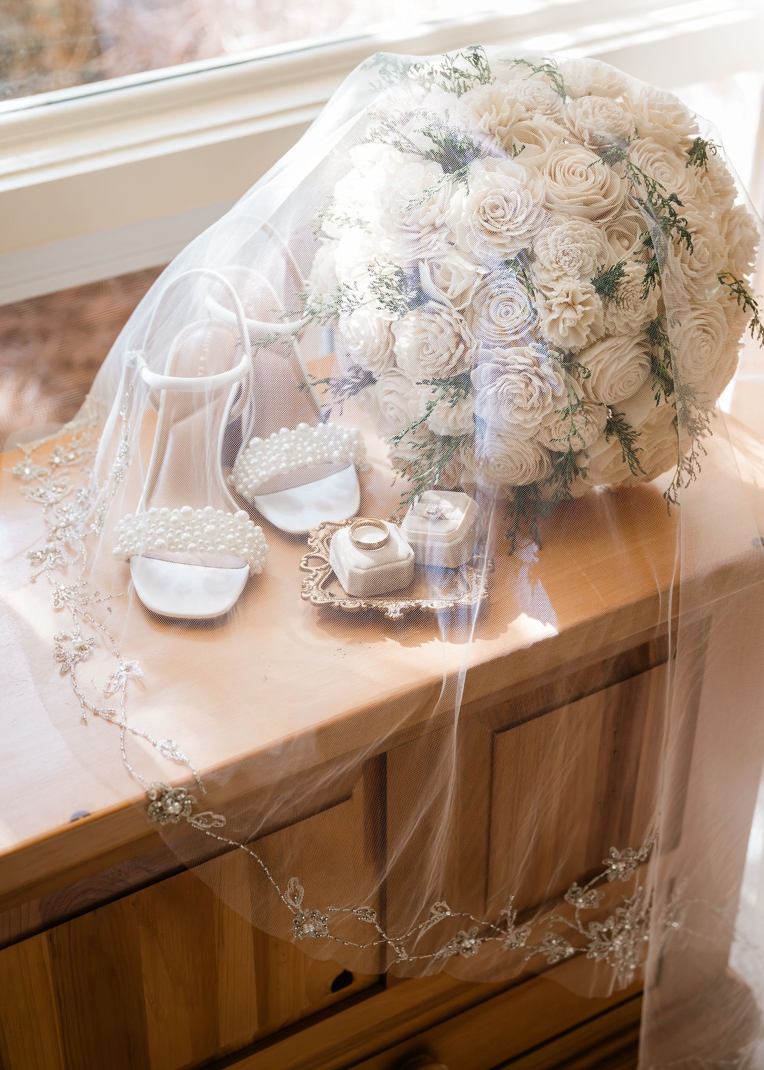 Elegant bridal flat lay featuring white rose bouquet, lace veil, pearl heels, and ring box on wooden dresser.