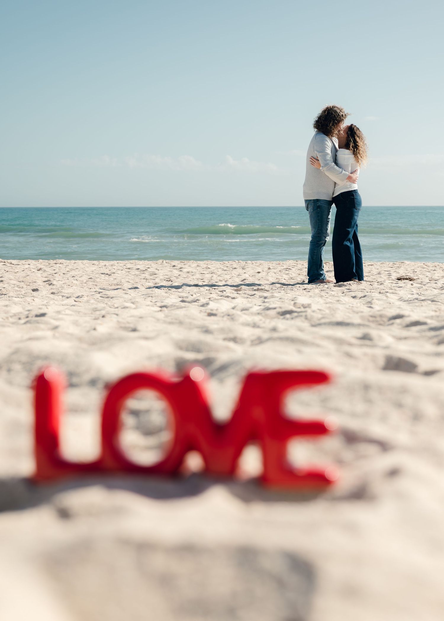 Red letters spelling out LOVE arranged on white beach sand with two people embracing in the background by the ocean.