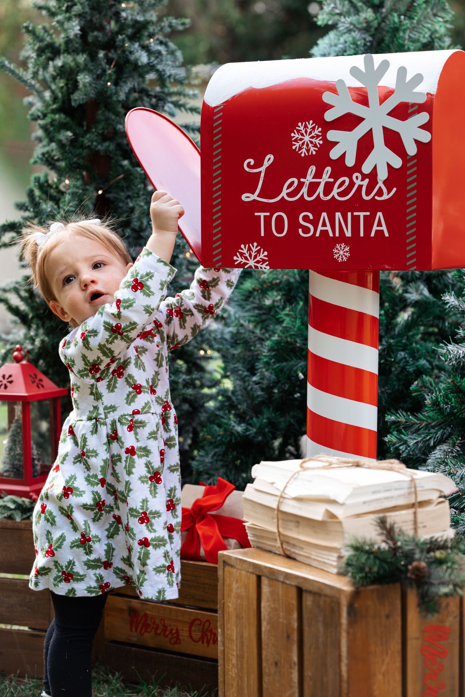 Child in holiday dress reaching for letters to Santa mailbox near Christmas trees.